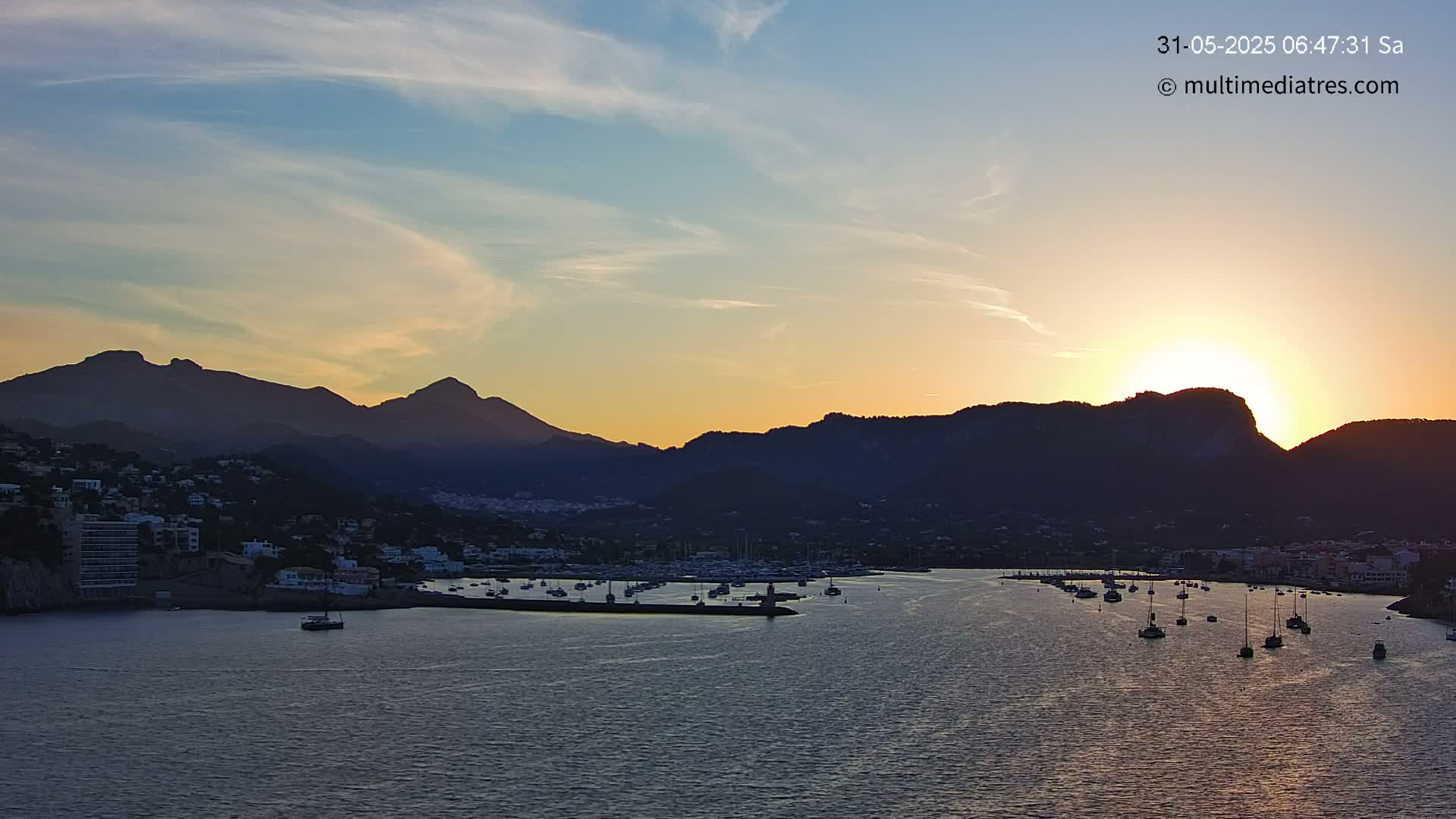 A coastal town nestled between mountains at sunset, with numerous boats in the harbor under a partly cloudy sky.