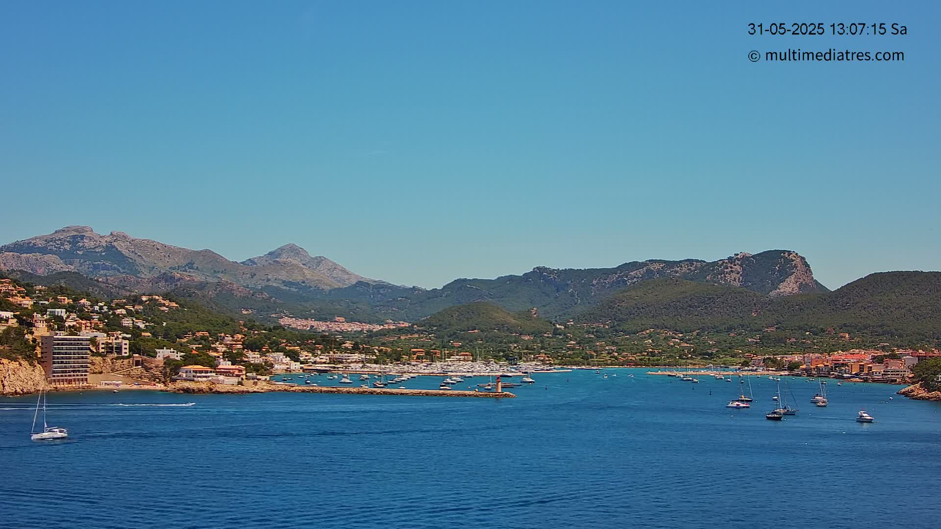 A picturesque coastal town nestled against a backdrop of mountains under a clear blue sky, with numerous boats in the calm bay.