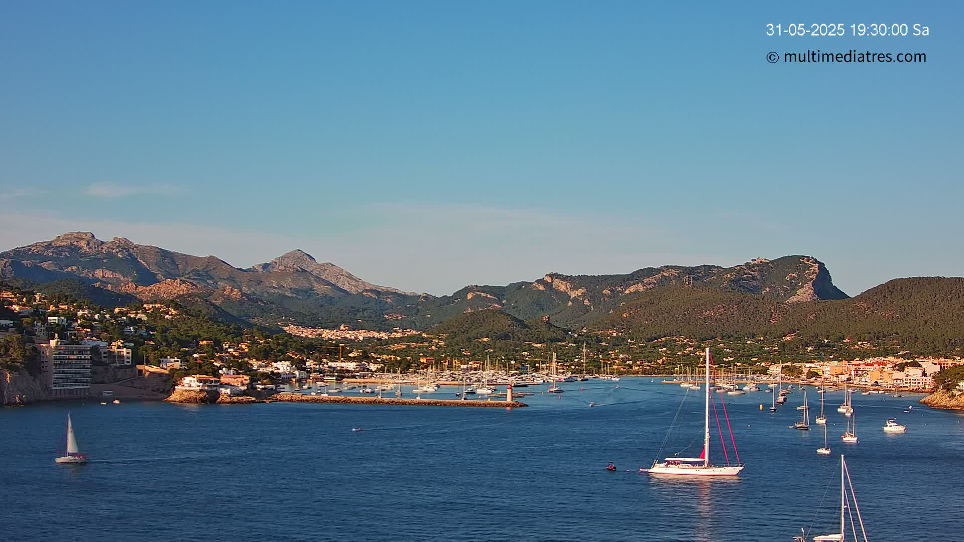 A calm, clear day shows a harbor filled with sailboats nestled between a hillside town and a mountain range.