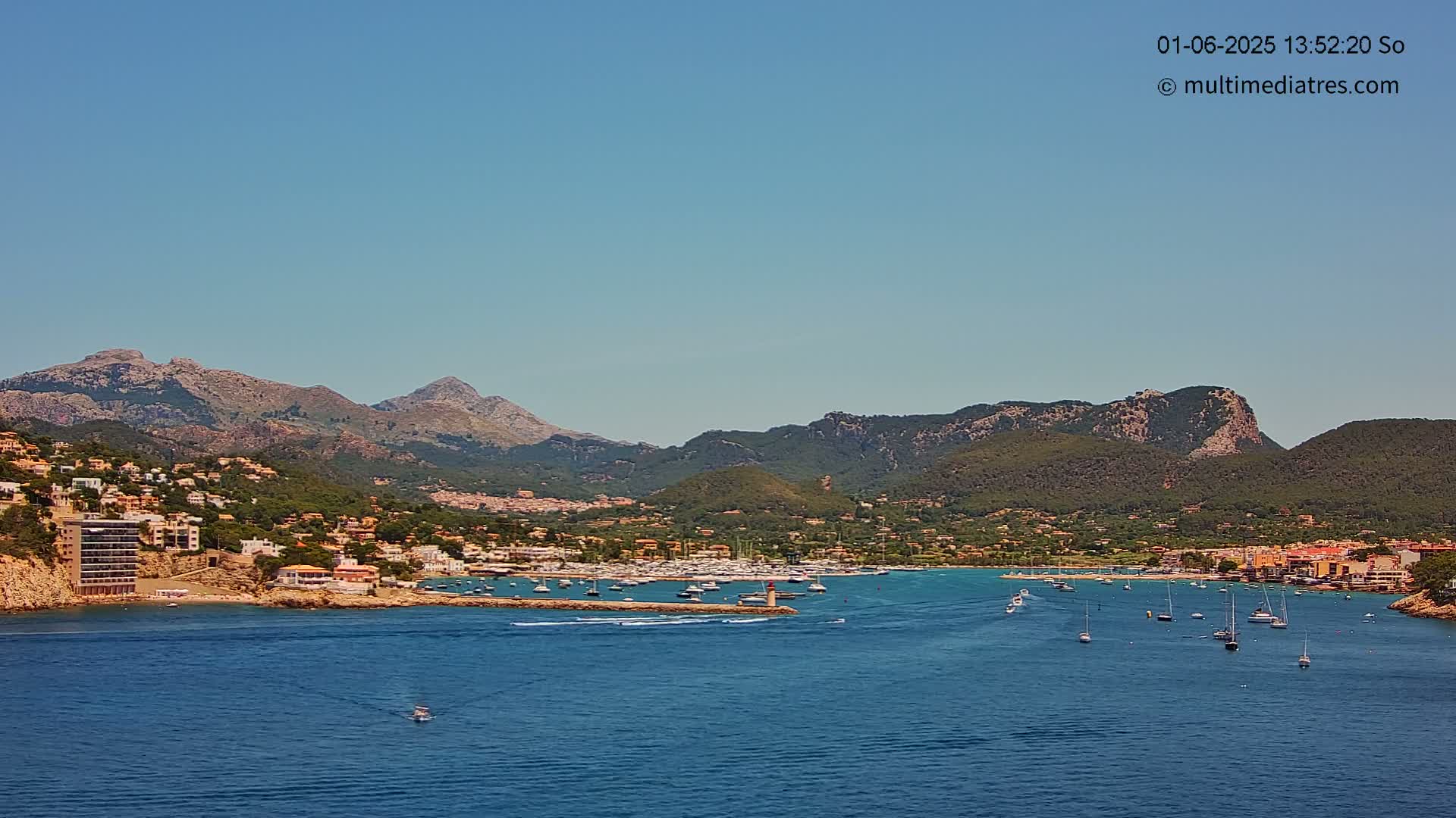 Under a clear blue sky, a calm bay filled with boats is nestled between a hillside town and a mountain range.