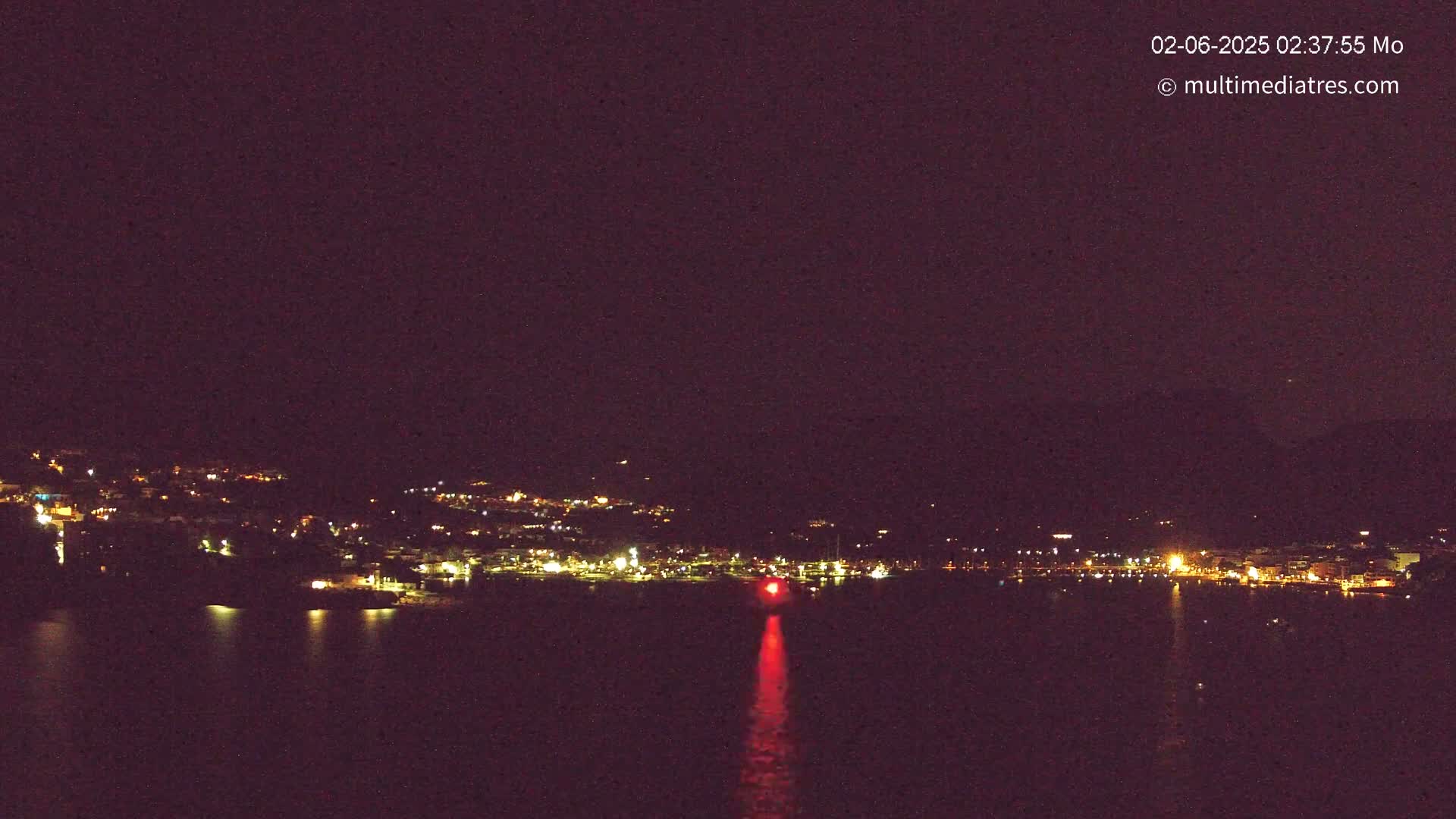 A nighttime view of a town's lights across a dark body of water, with a single red light visible on the water's surface.