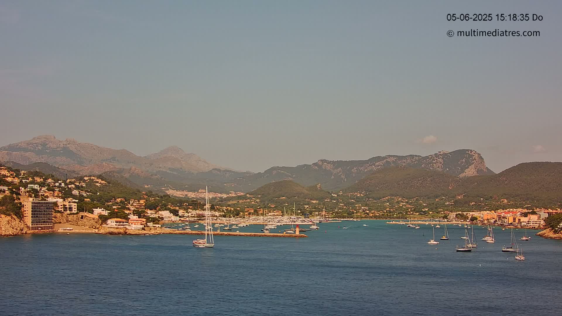 A hazy sunny day shows a calm bay filled with sailboats, a small marina, and a hillside town nestled against a mountain range.