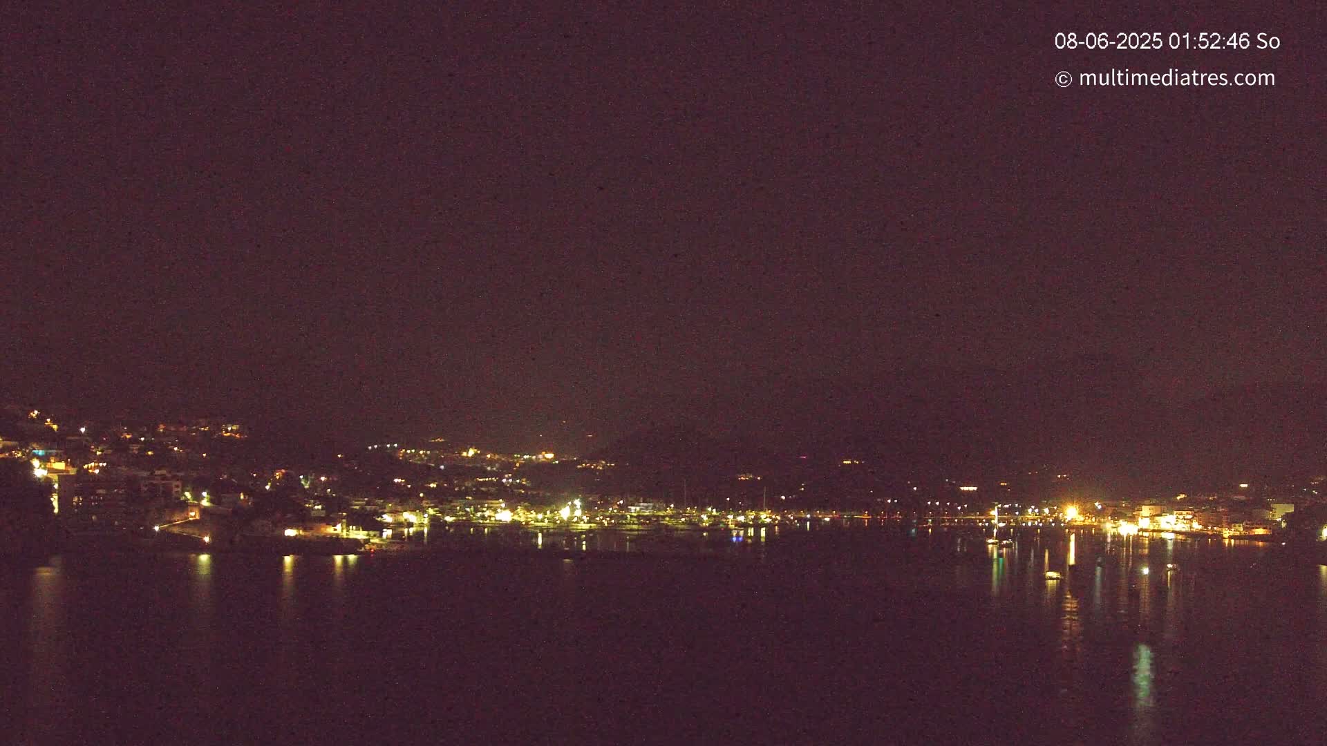 A nighttime view of a town situated on a hillside, with its lights reflecting on a calm body of water under a dark sky.