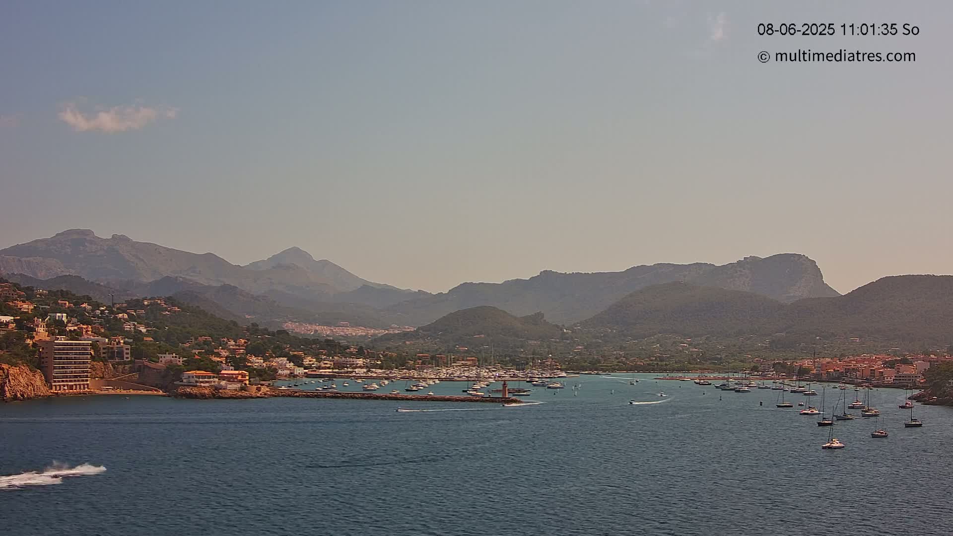 A hazy, sunny day reveals a coastal town nestled between mountains and a calm bay filled with numerous boats.