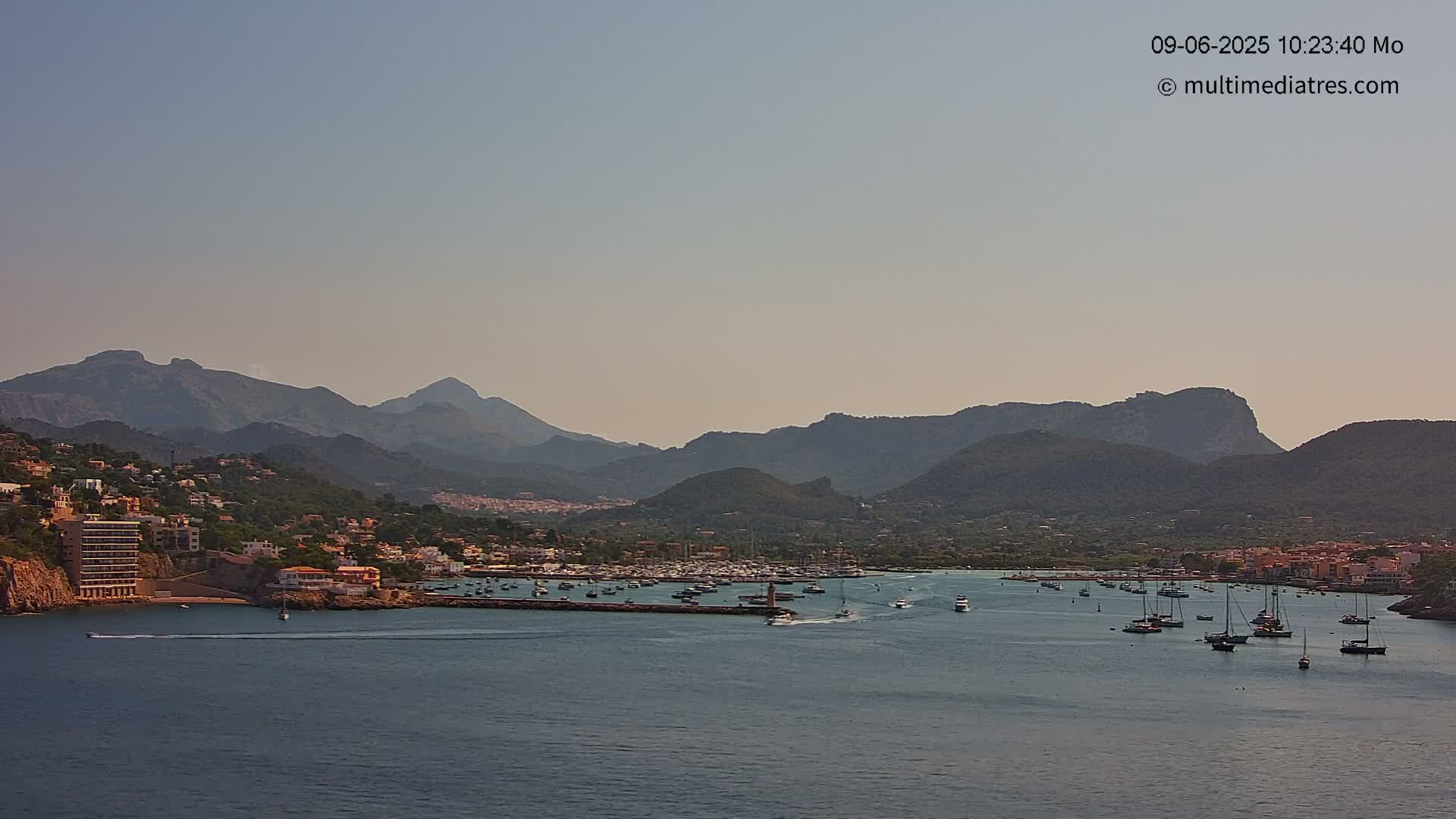 A harbor filled with boats is nestled in a coastal town, backed by a range of mountains under a hazy sky.