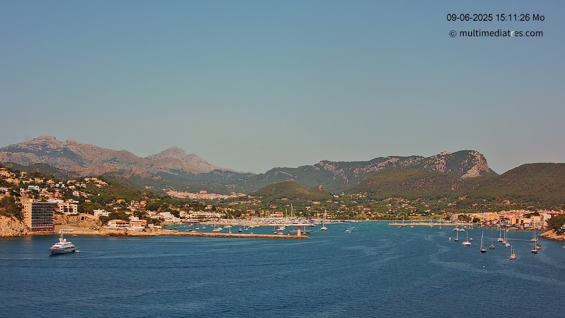 A yacht is in the foreground of a calm, blue bay, with a town nestled against mountains under a clear, sunny sky.