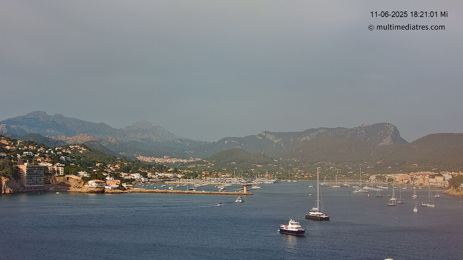 A hazy coastal town nestled at the base of mountains, with a calm harbor filled with boats.