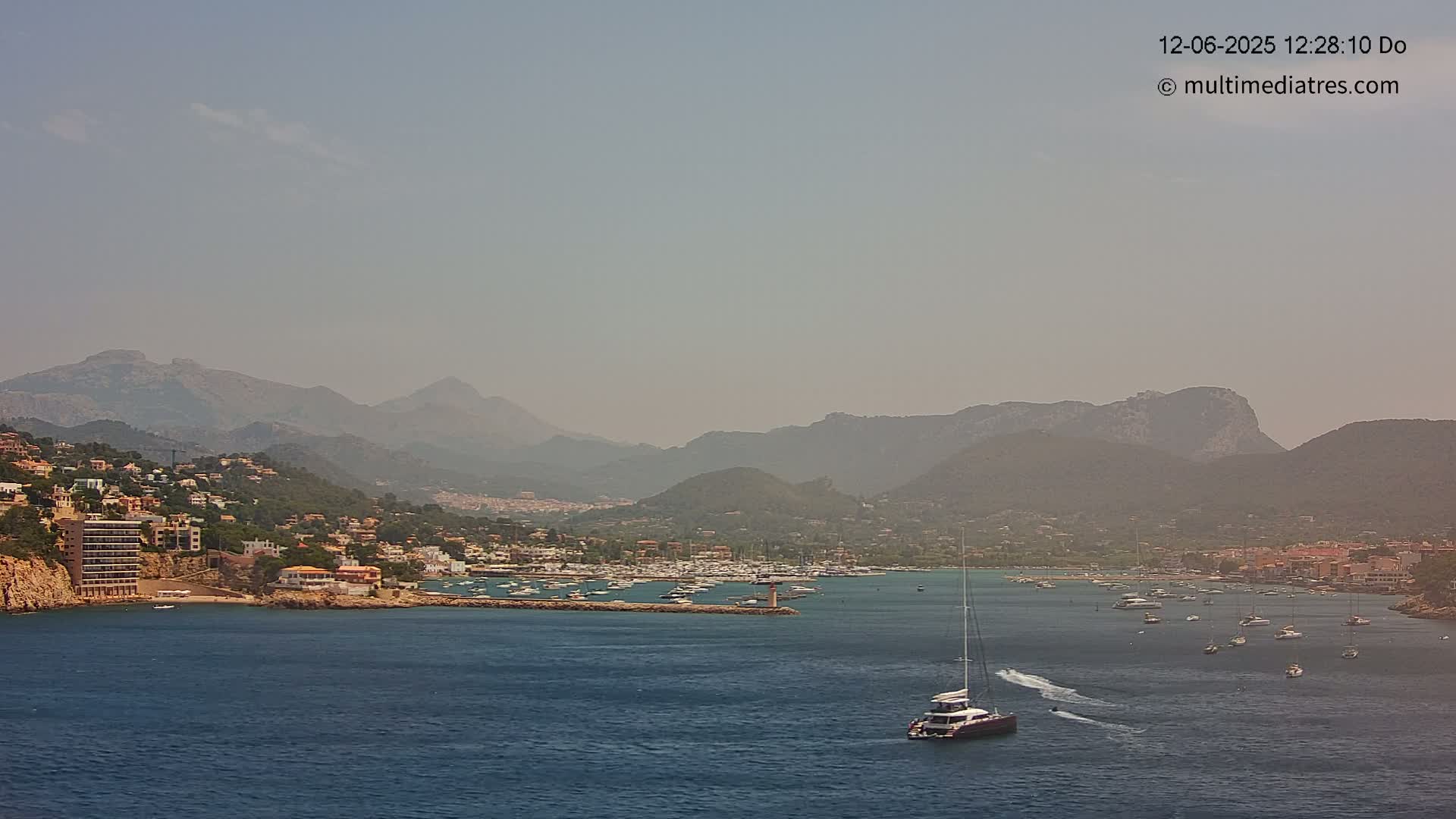 A hazy, sunny day reveals a coastal town nestled between mountains and a calm bay filled with numerous boats, one of which is a large catamaran.