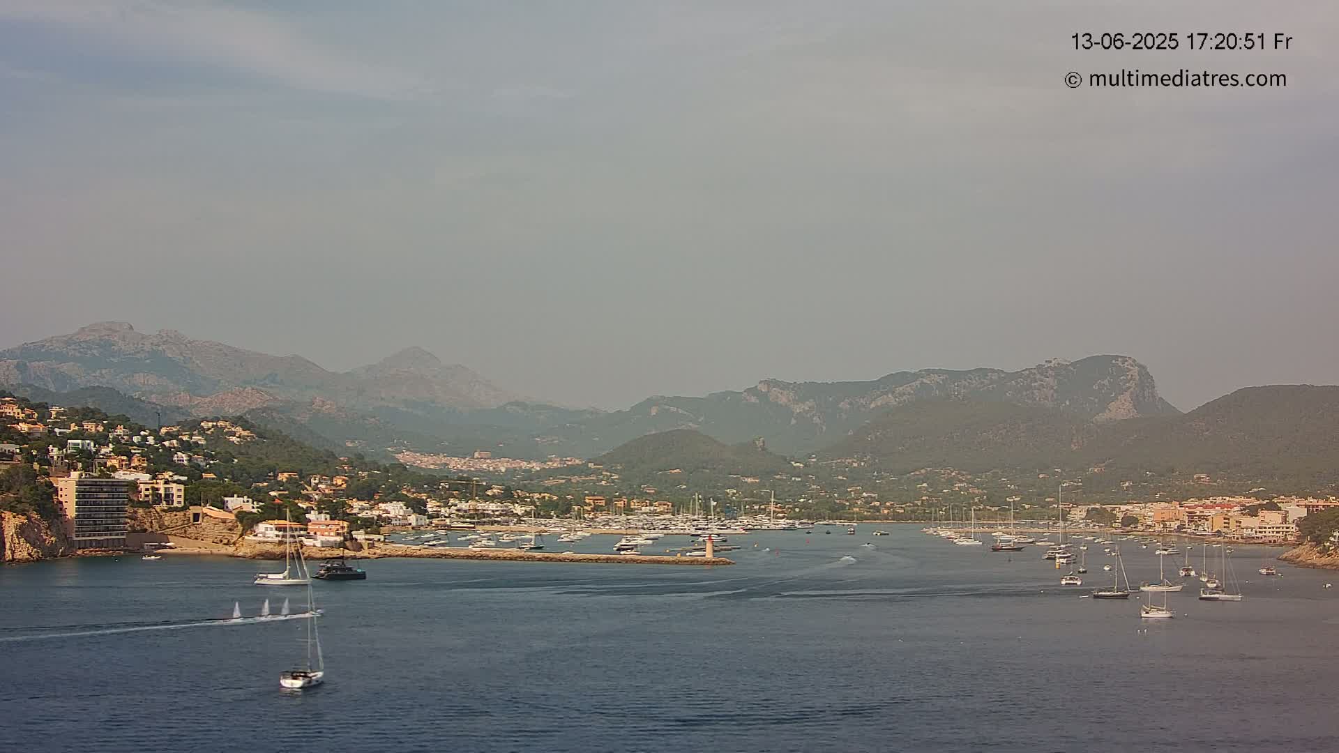 A hazy coastal town nestled at the base of mountains, with a harbor filled with boats, is visible under a pale sky.