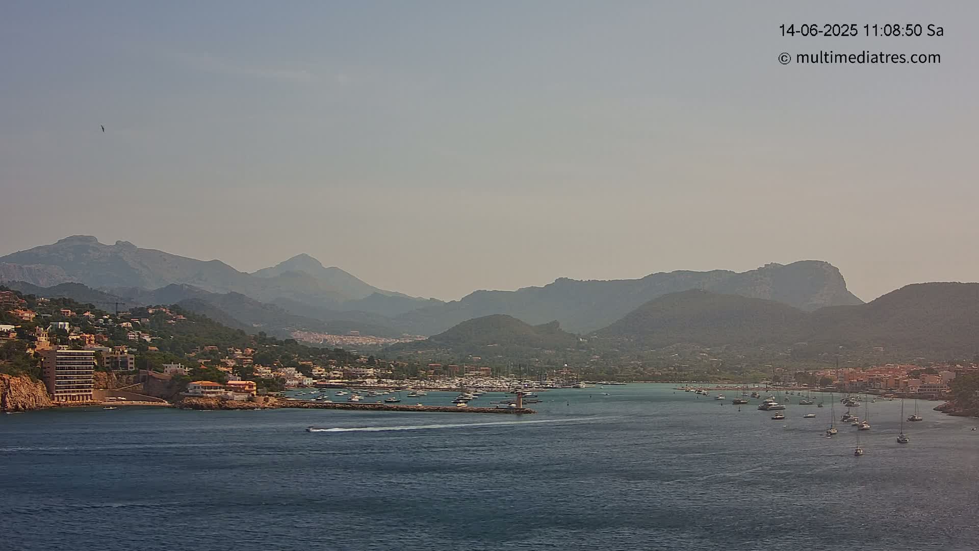A hazy, sunny day reveals a coastal town nestled between mountains and a calm bay filled with sailboats.