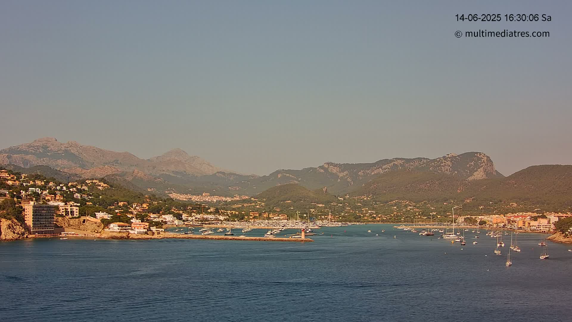 A hazy day reveals a coastal town nestled between a calm bay filled with boats and a range of mountains.