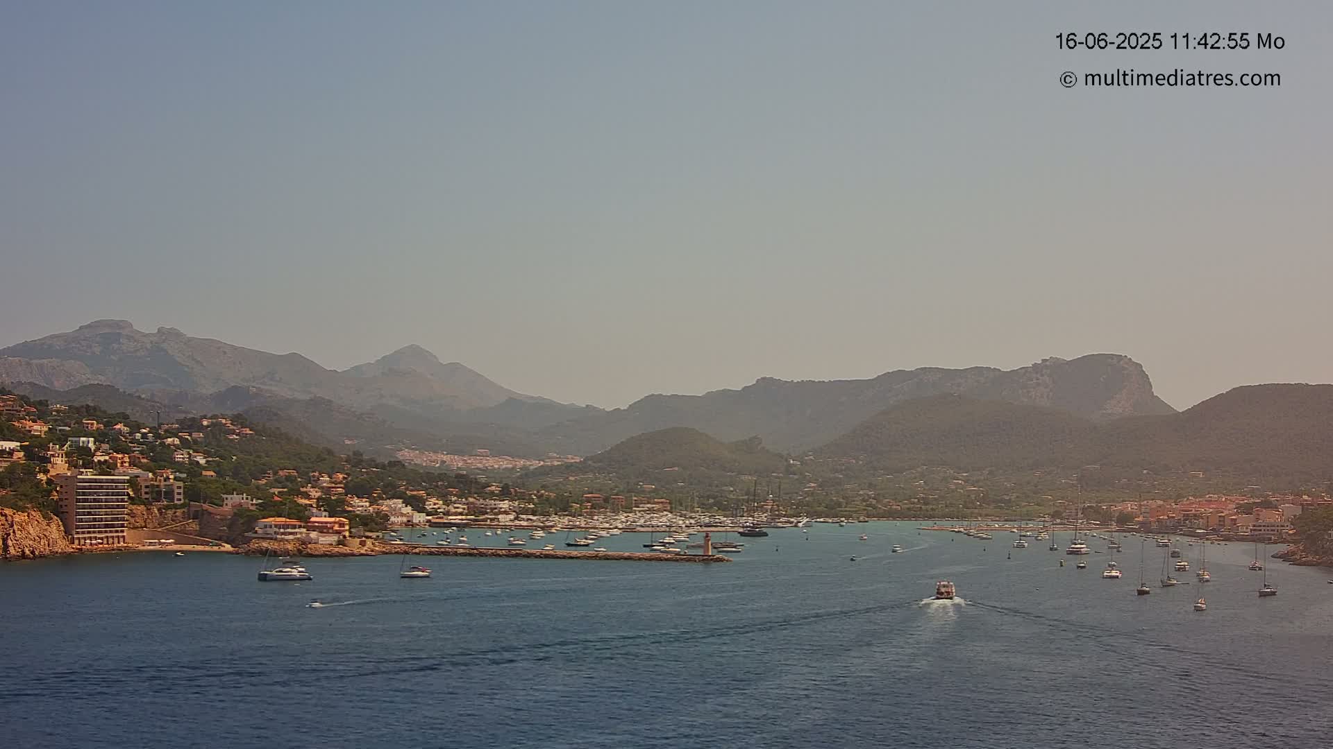 A hazy, sunny day shows a coastal town nestled between mountains and a calm bay filled with boats.