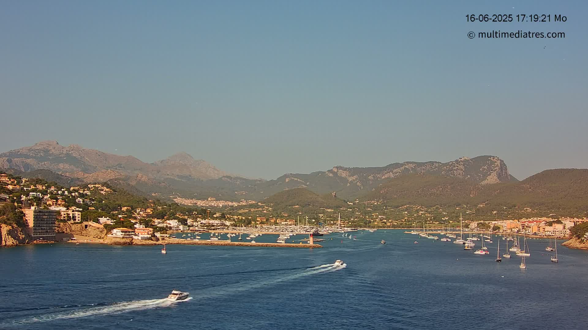 A harbor filled with boats is nestled between a coastal town and a mountain range under a clear, hazy sky.