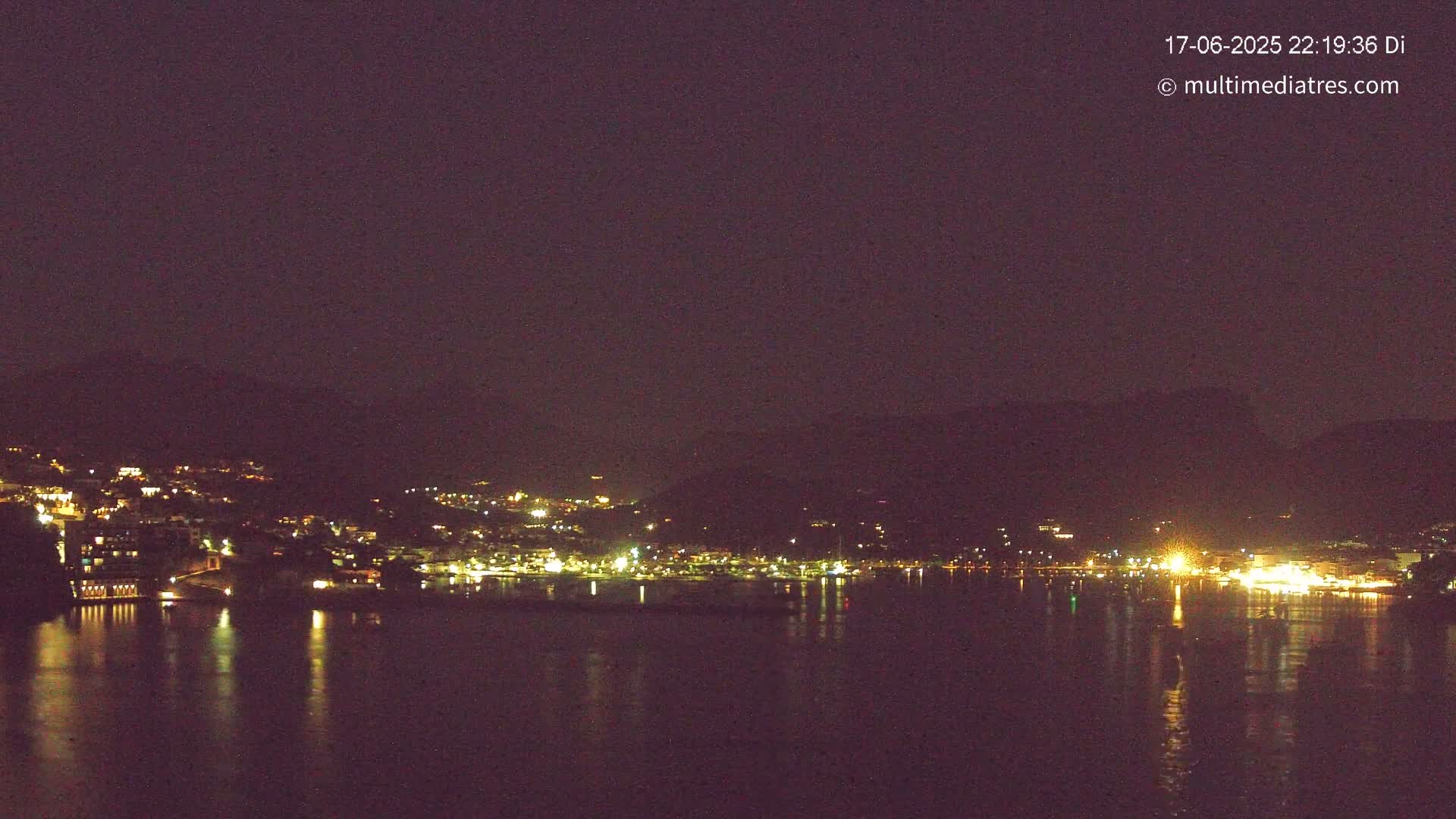 A nighttime view of a town situated on a body of water, nestled against dark hills under a clear, dark sky.