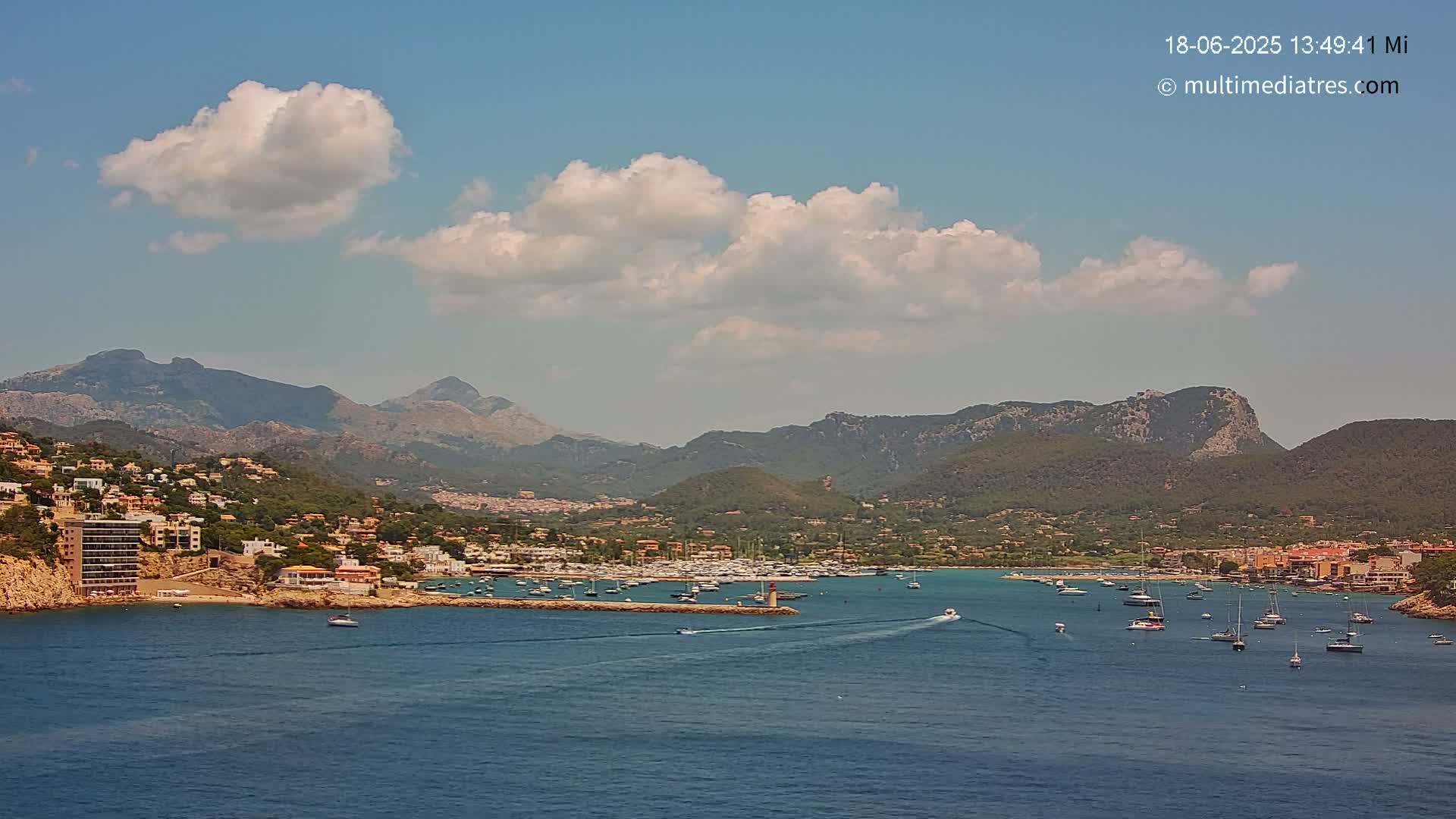 A harbor filled with boats is nestled between a hillside town and a mountain range under a partly cloudy sky.