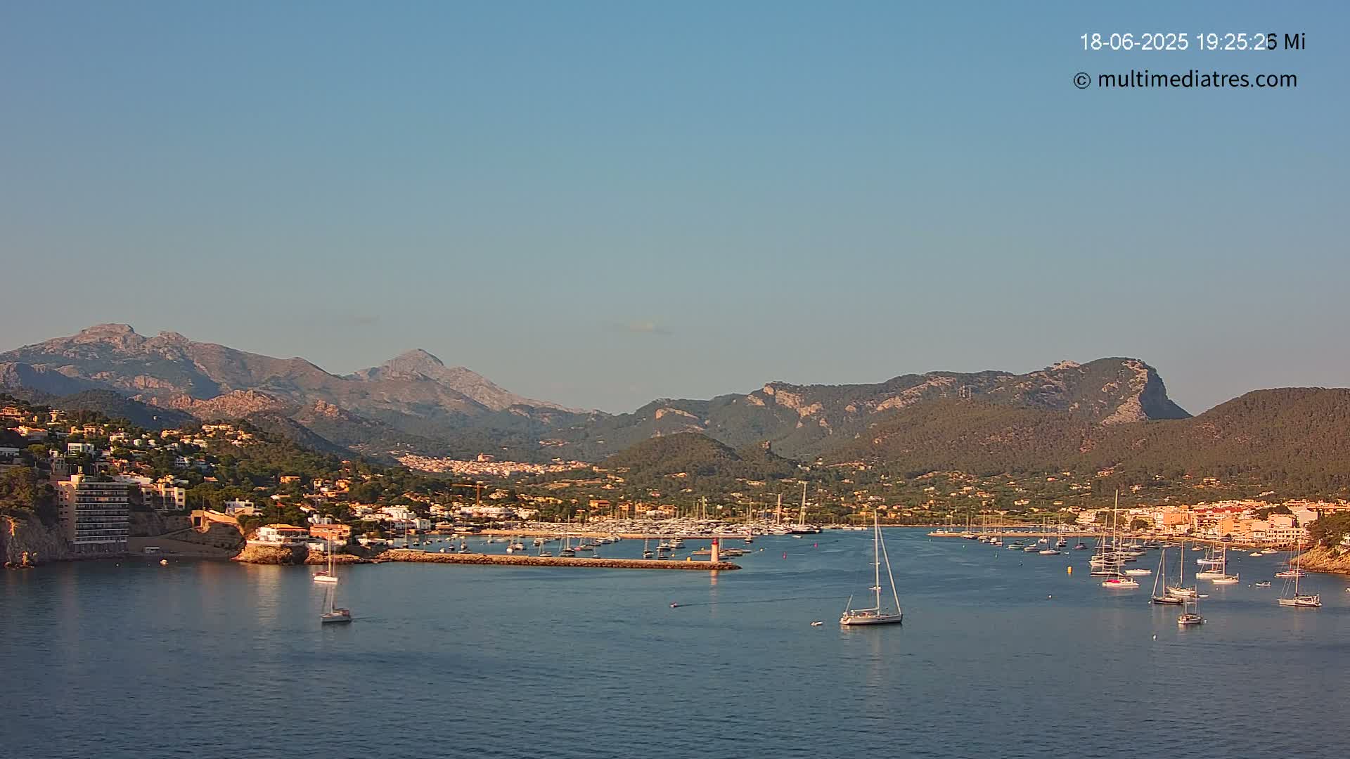 A clear, sunny day reveals a coastal town nestled between mountains and a calm bay filled with sailboats.