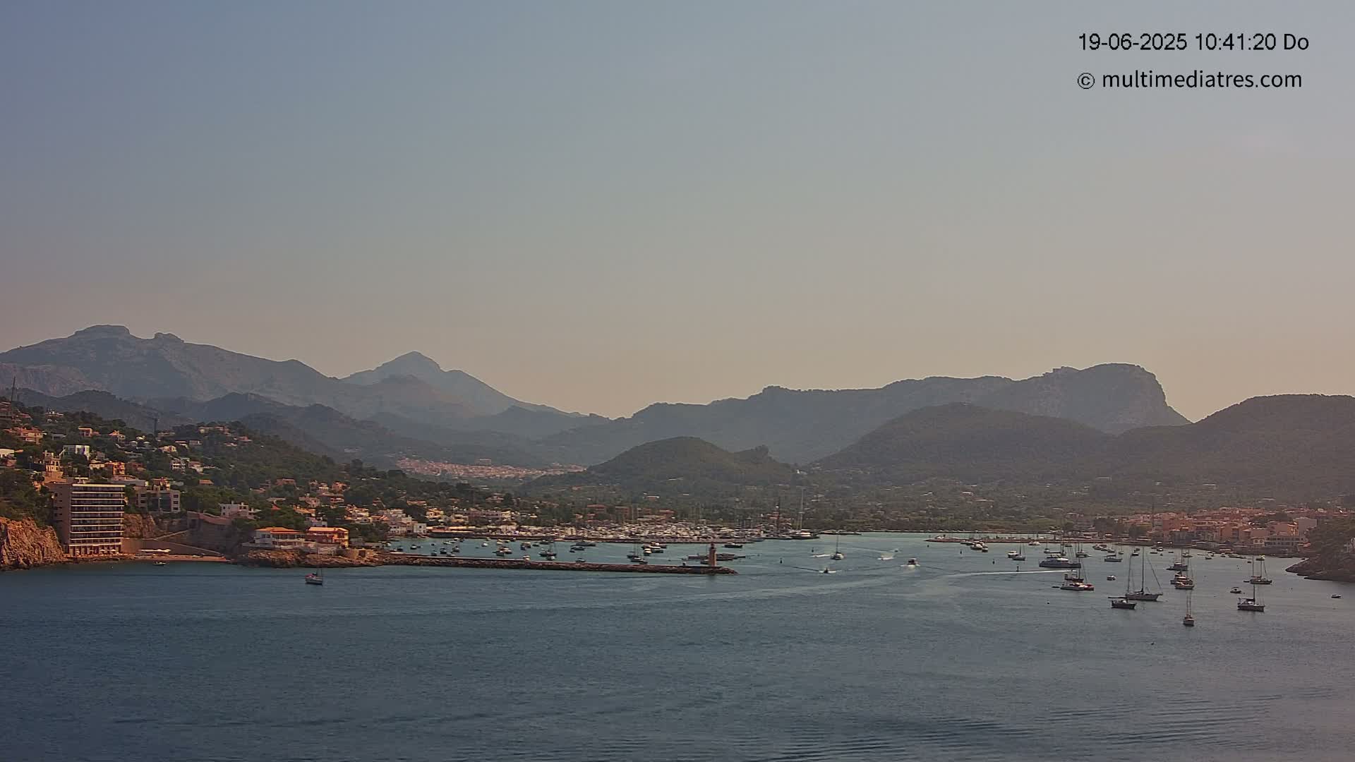 A hazy, sunny day reveals a harbor filled with boats nestled between a hillside town and a mountain range.