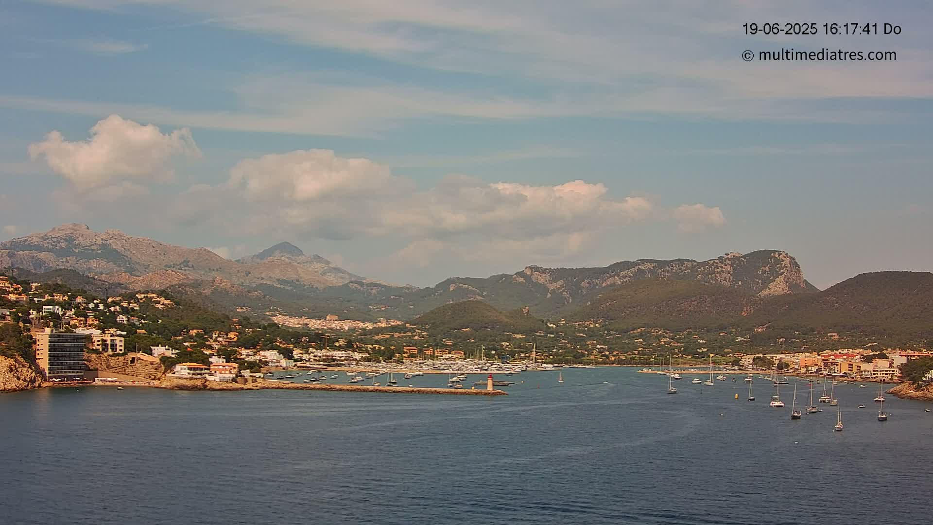 A partly cloudy sky overlooks a harbor filled with sailboats nestled between a coastal town and a mountainous backdrop.