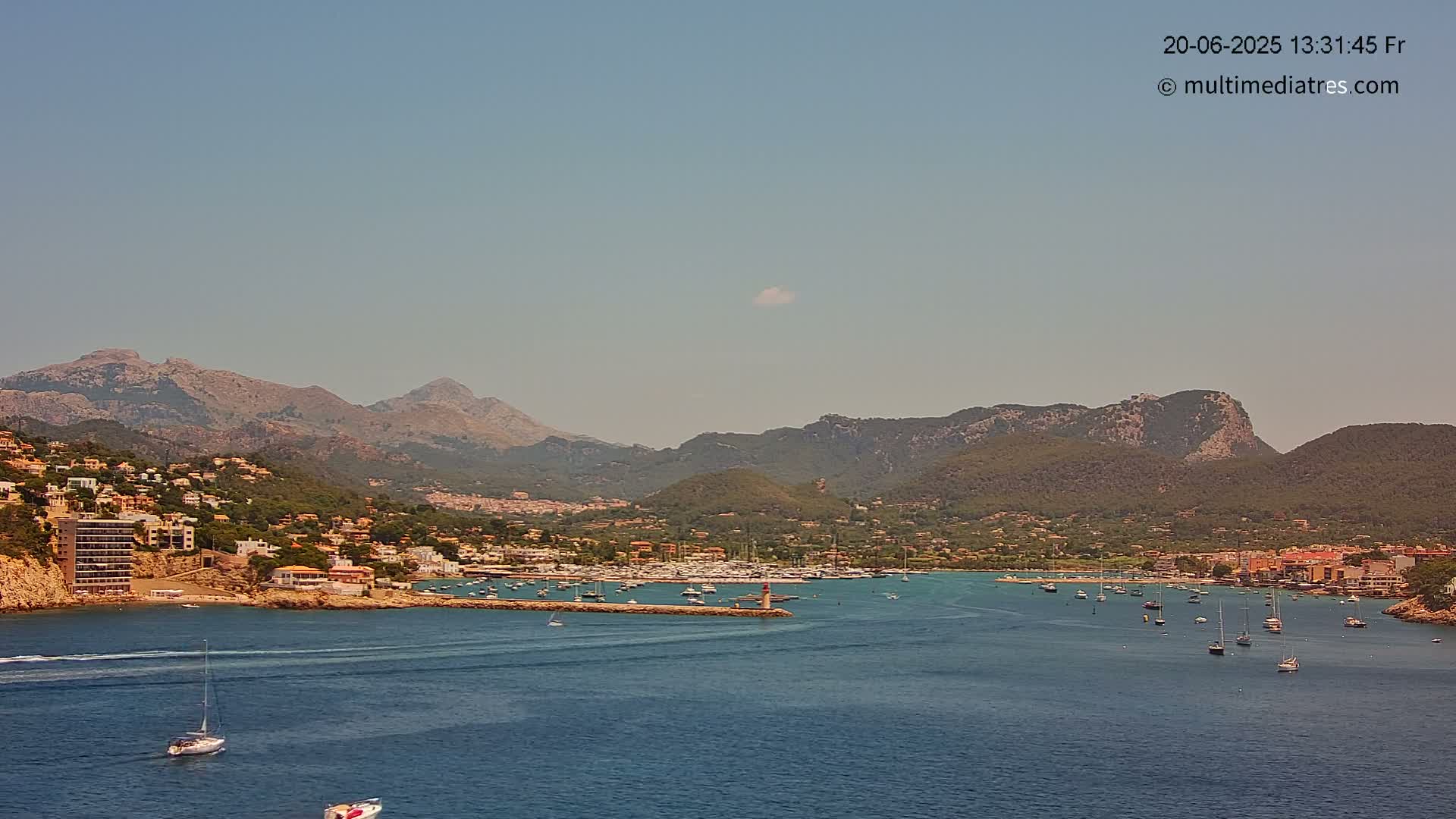 A harbor filled with sailboats sits nestled among a hillside town, all backed by a range of mountains under a clear, sunny sky.