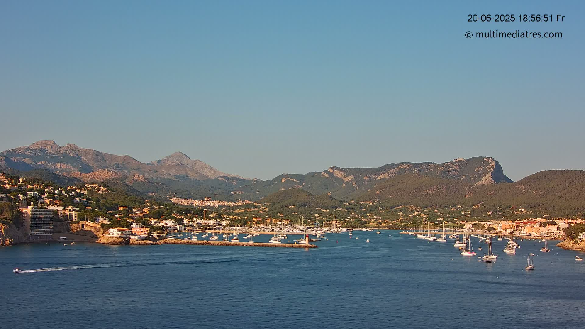 A harbor filled with sailboats is nestled between a coastal town and mountains under a clear, bright sky.