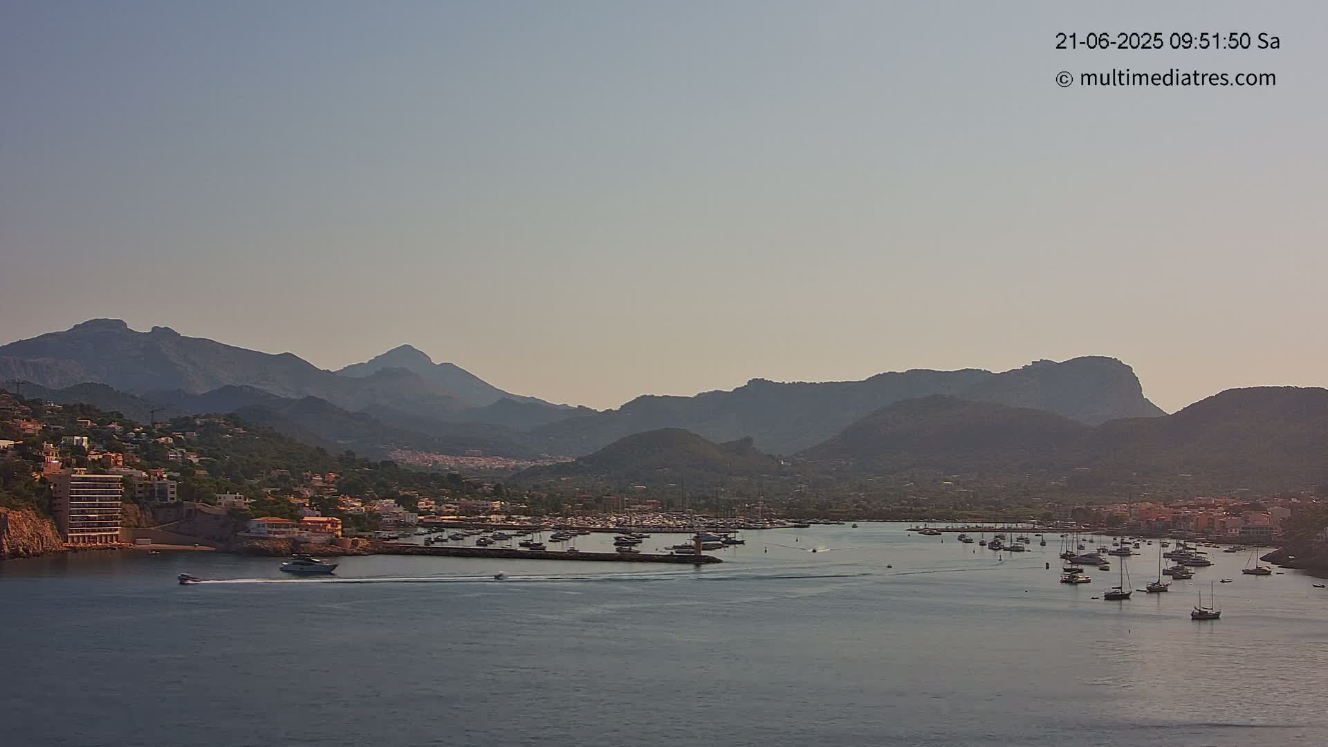 A calm harbor filled with boats sits nestled between a coastal town and a range of hazy mountains under a clear sky.
