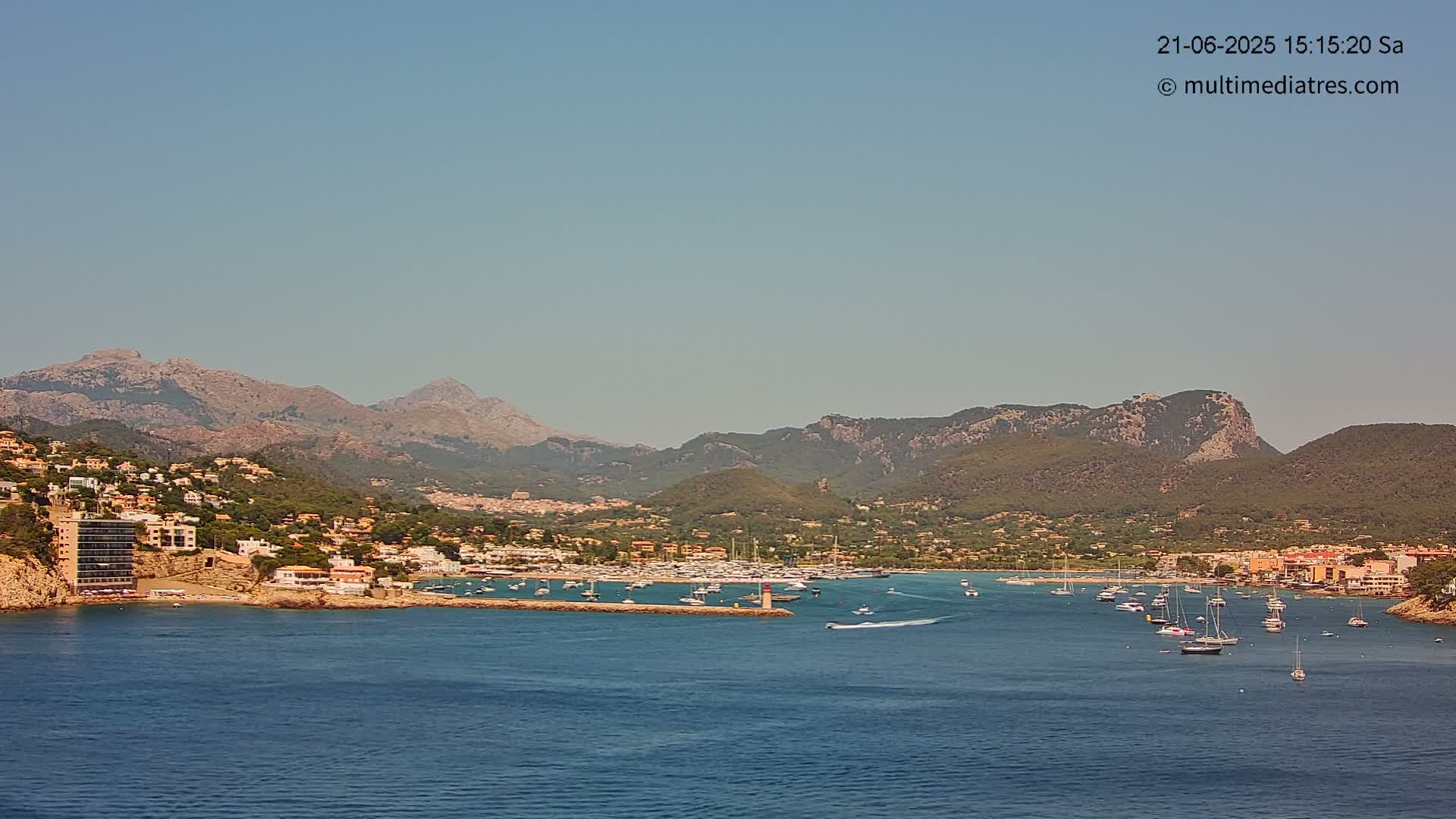 A clear, sunny day shows a coastal town nestled between mountains and a calm bay filled with boats.