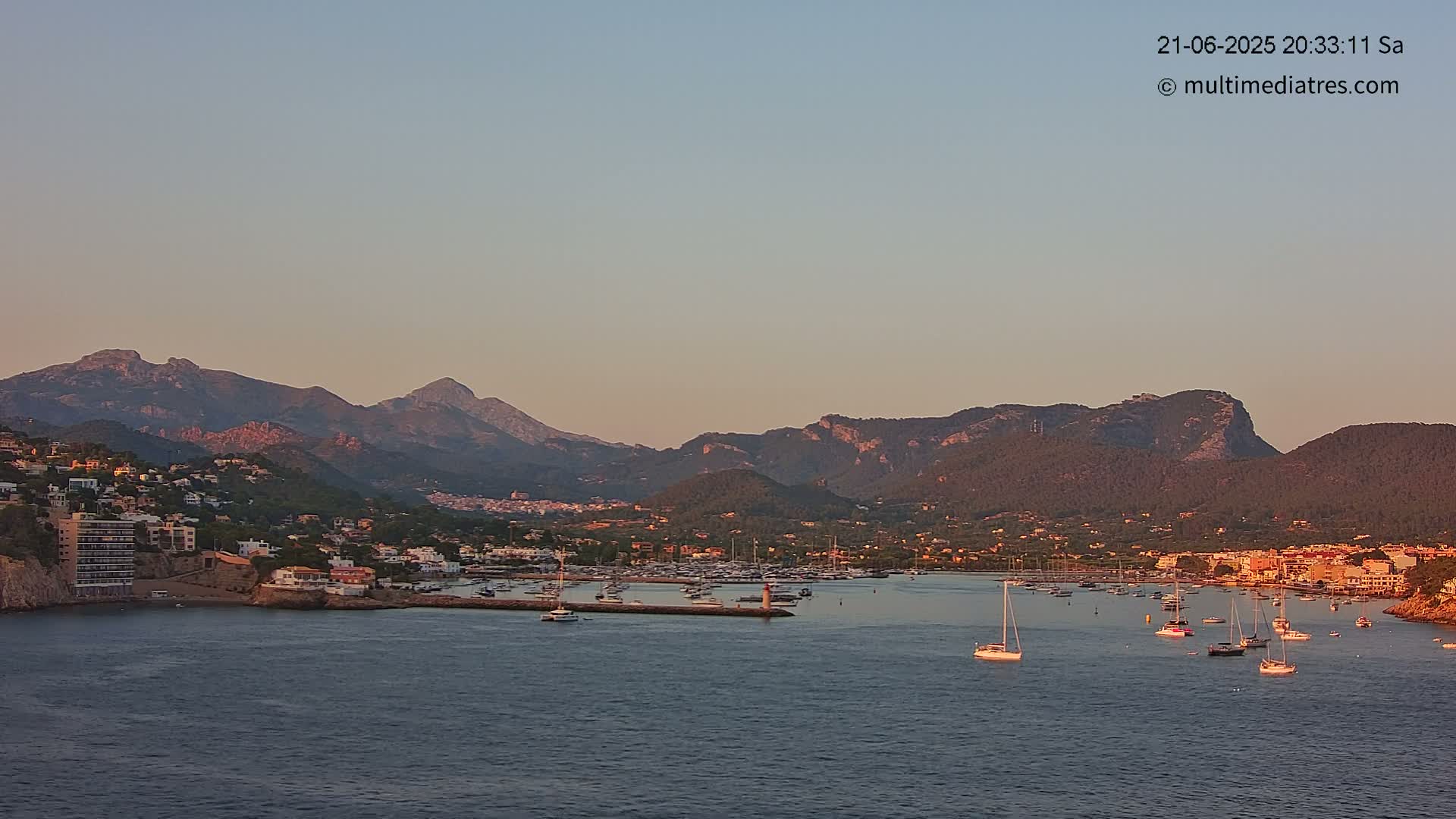A calm harbor filled with sailboats sits nestled in a coastal town at the base of a mountain range under a pale sunset sky.
