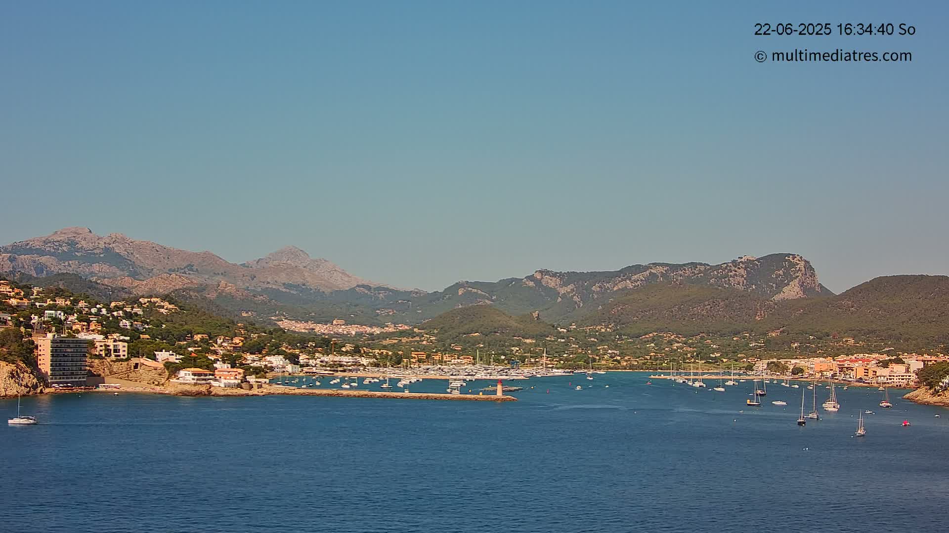 A coastal town nestled in a bay, surrounded by mountains under a clear, sunny sky, with numerous boats in the harbor.