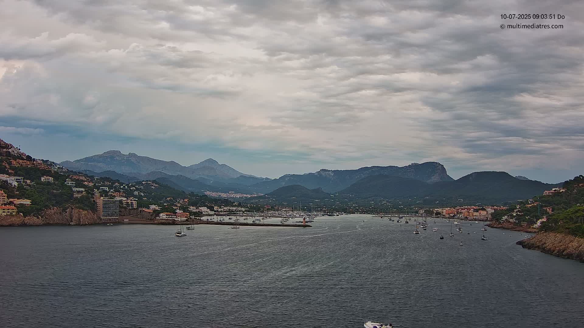 A harbor filled with boats is nestled between hills and mountains under a cloudy sky.