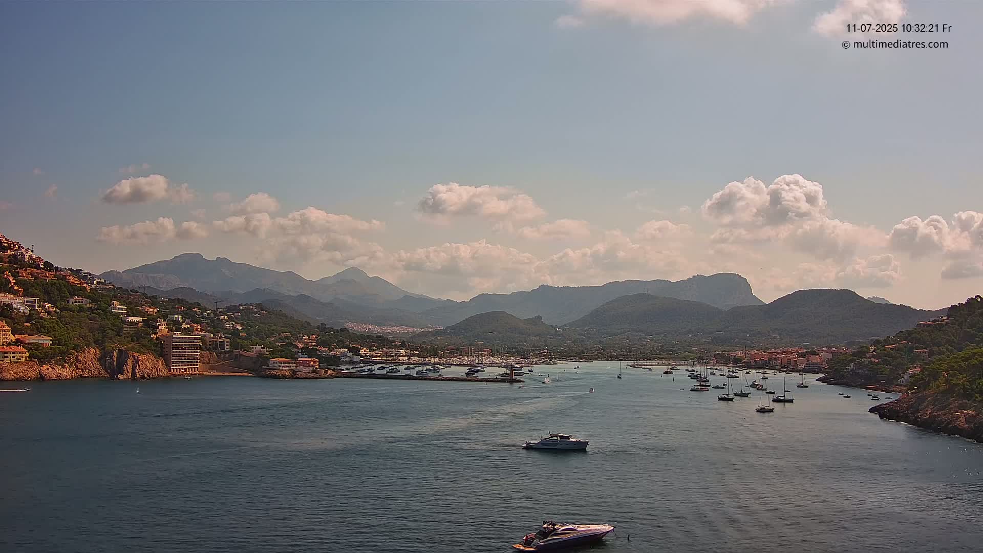 A partly cloudy day shows a harbor filled with boats, nestled between hillsides covered in buildings and lush greenery, all set against a backdrop of mountains.