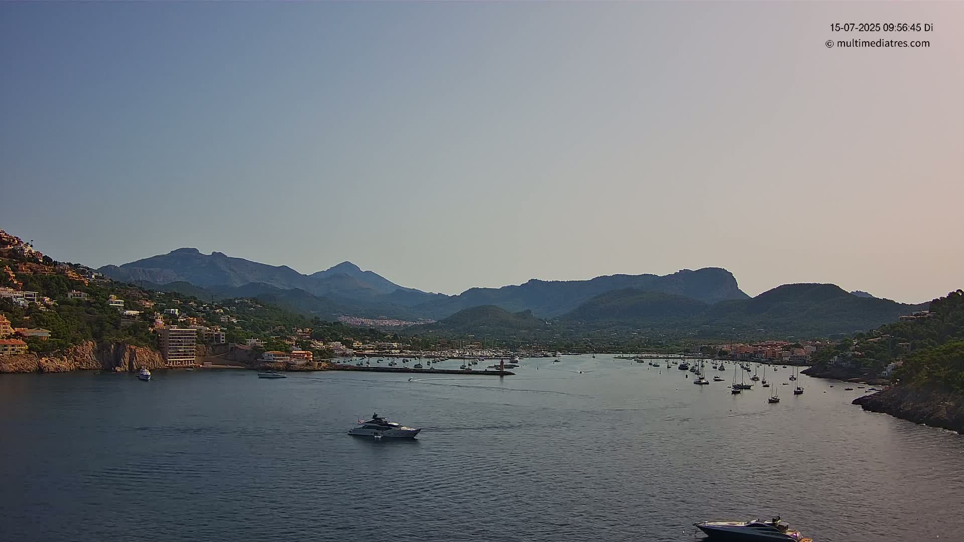 A calm bay with numerous boats is nestled between a hillside town and a mountain range under a clear sky.