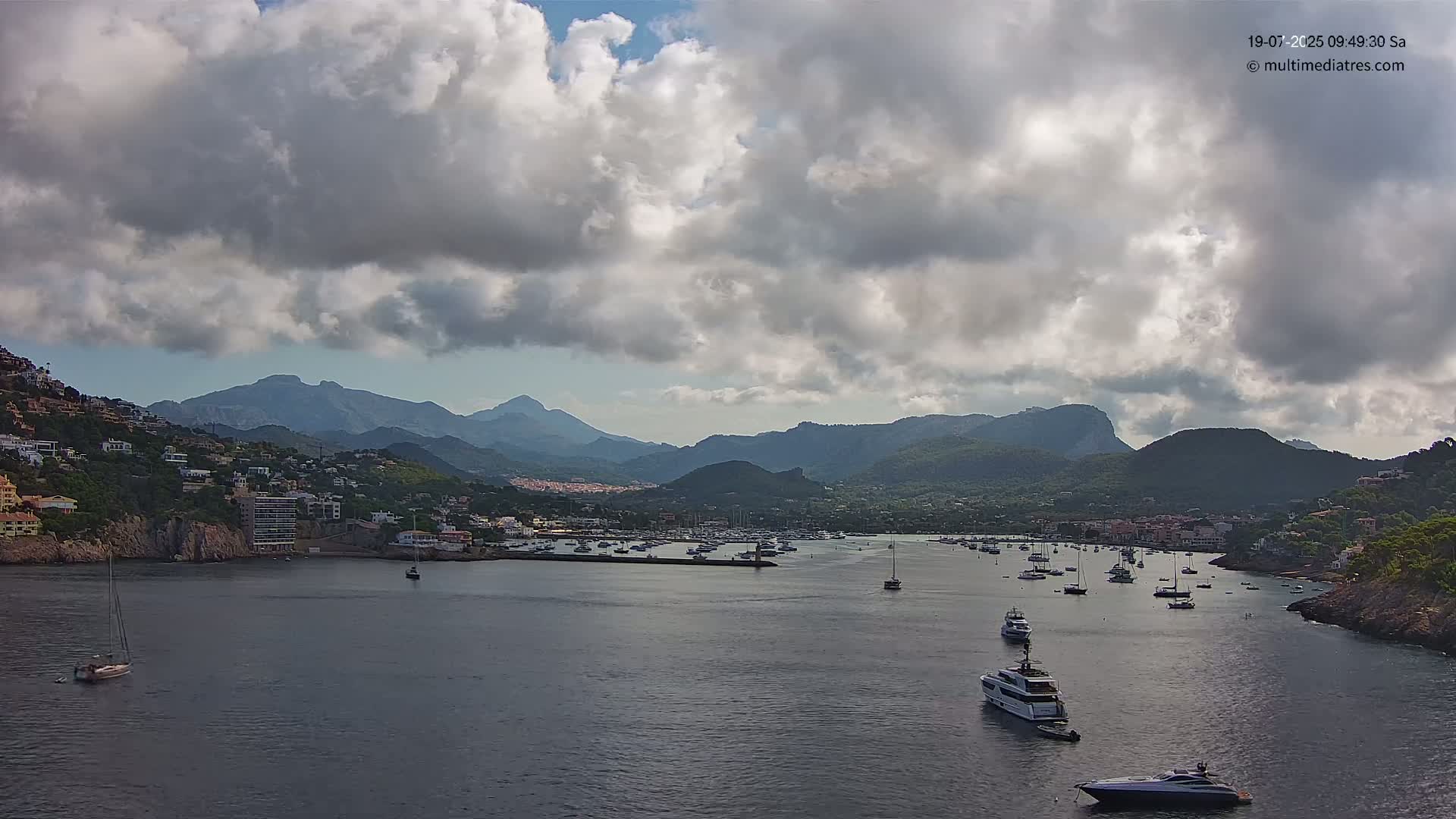 A calm bay filled with boats is nestled between hillsides dotted with buildings, all under a partly cloudy sky.