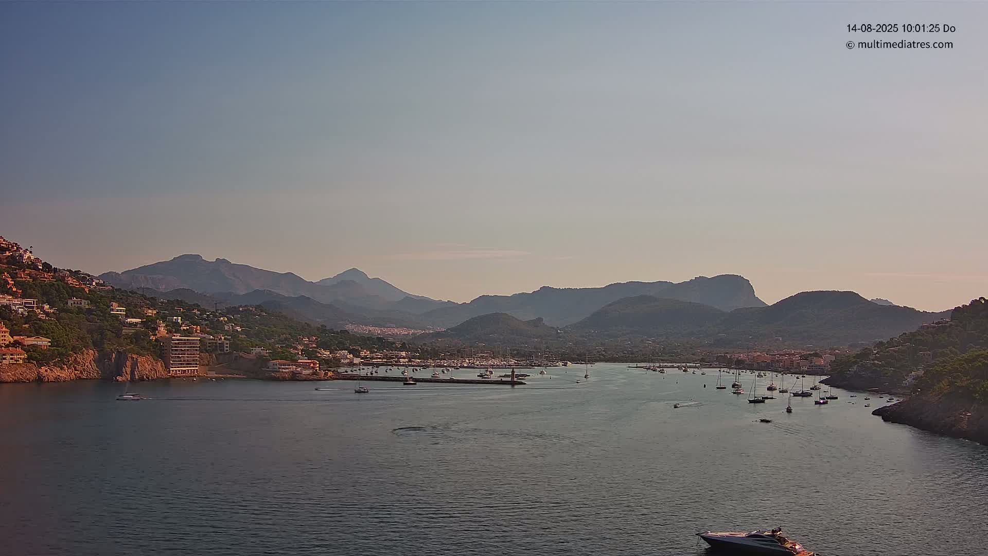 A calm body of water filled with boats is nestled between hillsides dotted with buildings, all under a clear sky.