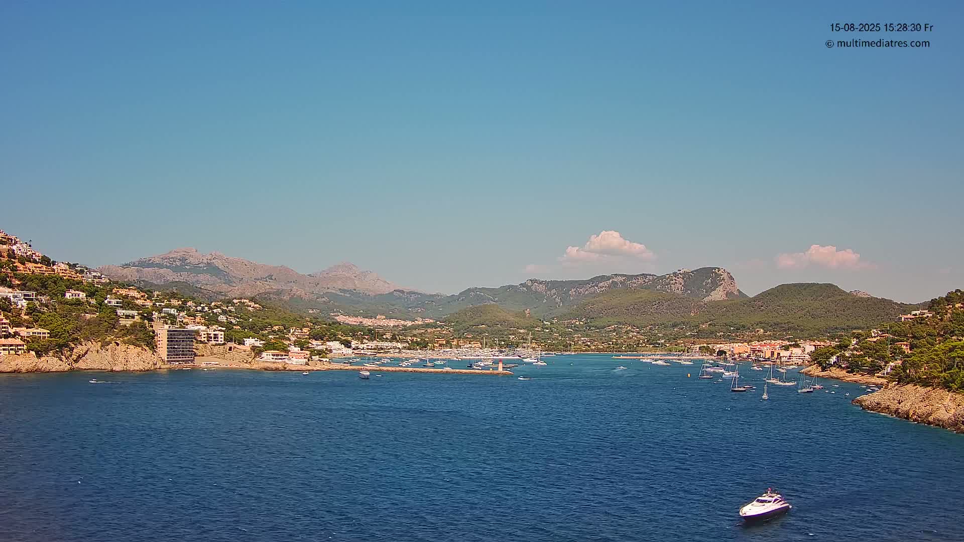 A calm body of water filled with boats is nestled between hillsides dotted with buildings, all under a clear sky.