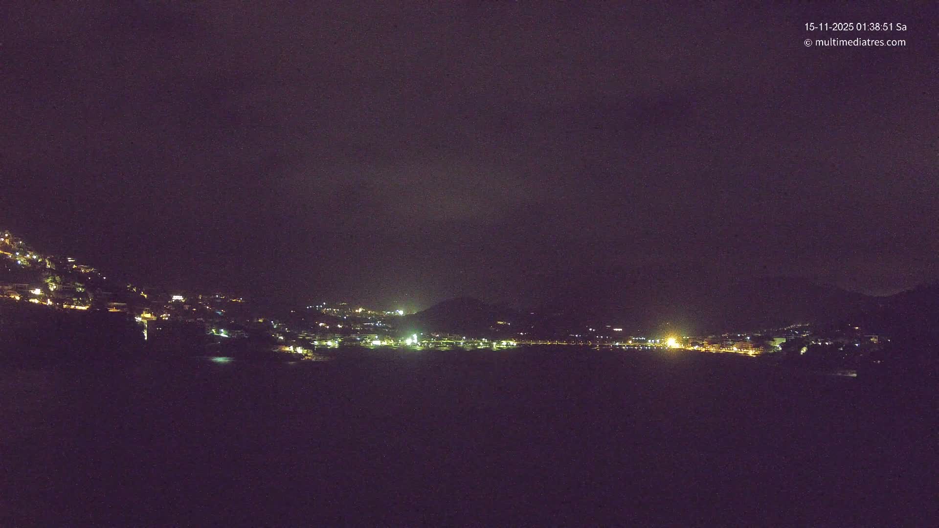 A clear night view captures a brightly lit coastal town nestled against dark mountains, with numerous lights reflecting on the calm water.