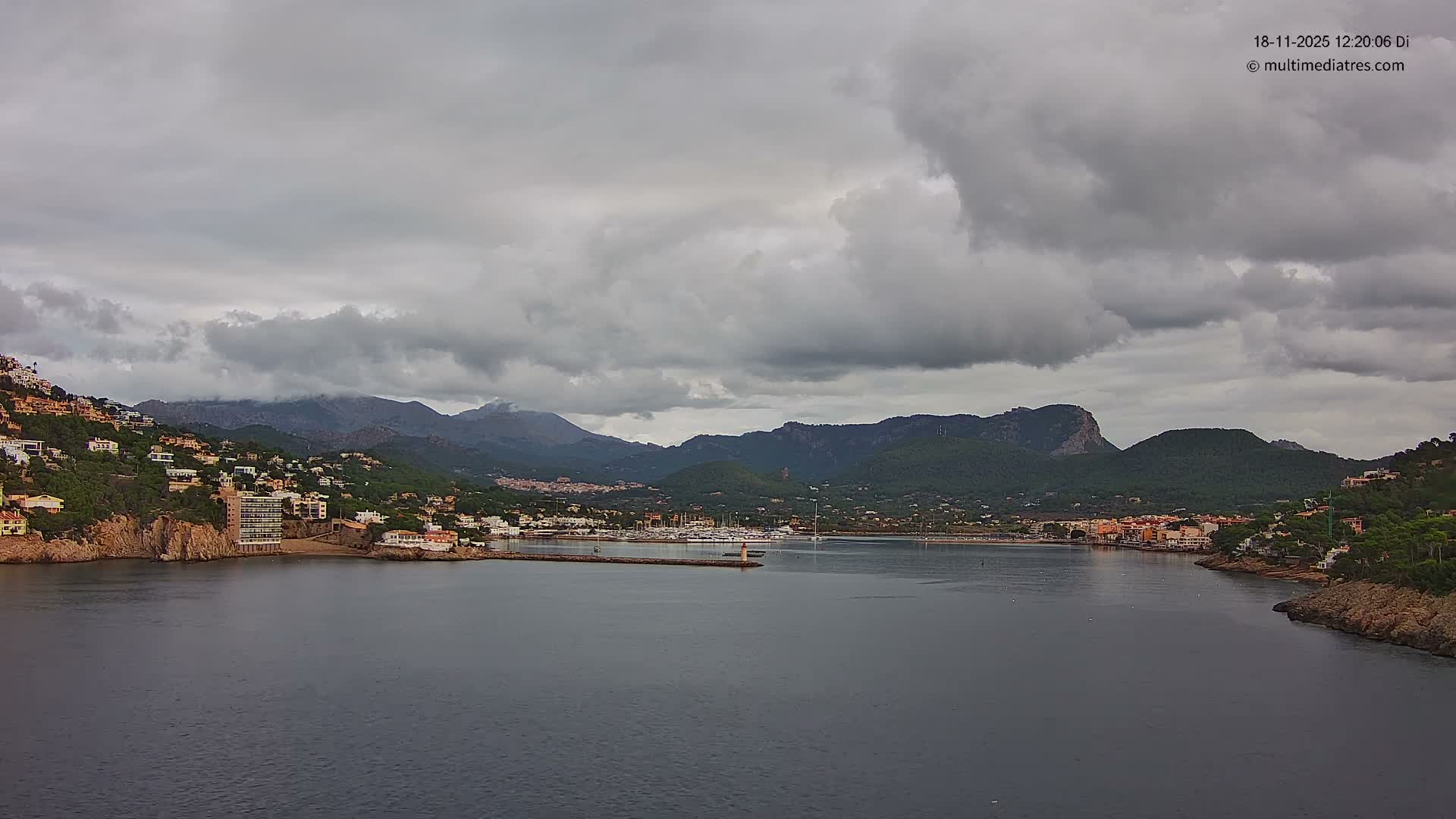 A clear night view captures a brightly lit coastal town nestled against dark mountains, with numerous lights reflecting on the calm water.