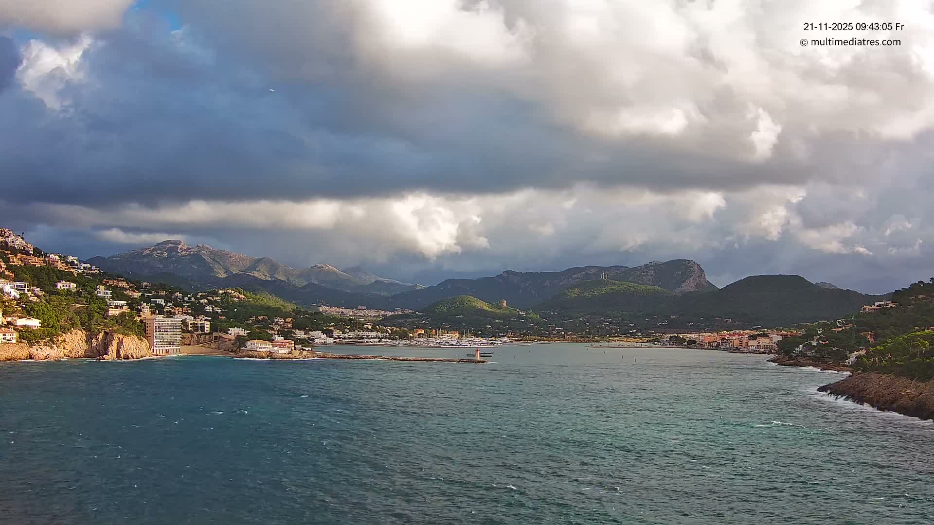 A coastal town with a marina and hillside homes is set against rugged mountains, with choppy waters in the foreground under a heavily overcast sky broken by dramatic shafts of sunlight.