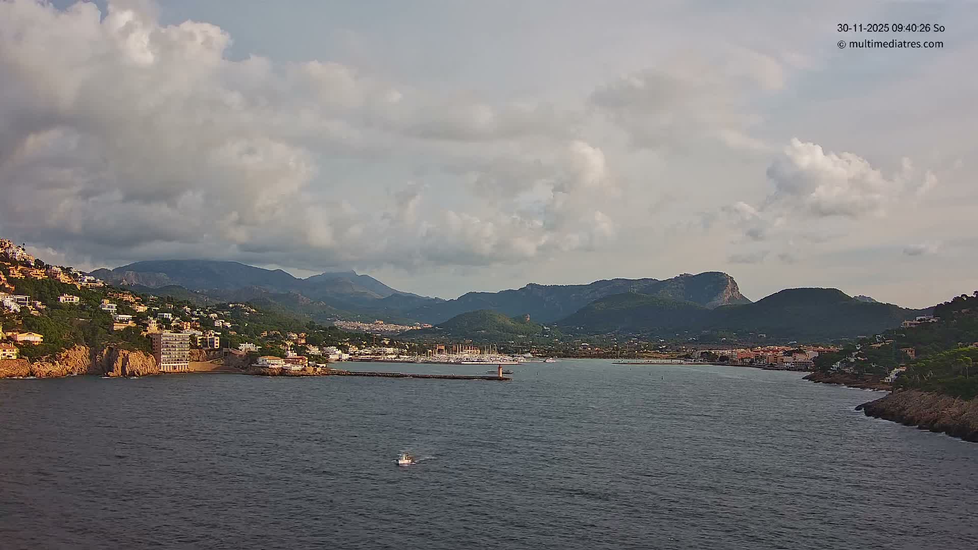 Under a heavily overcast sky, a coastal town with buildings on green hillsides encircles a bay dotted with a marina and a lighthouse on a breakwater, while a small boat cruises the dark water against a backdrop of distant mountains.