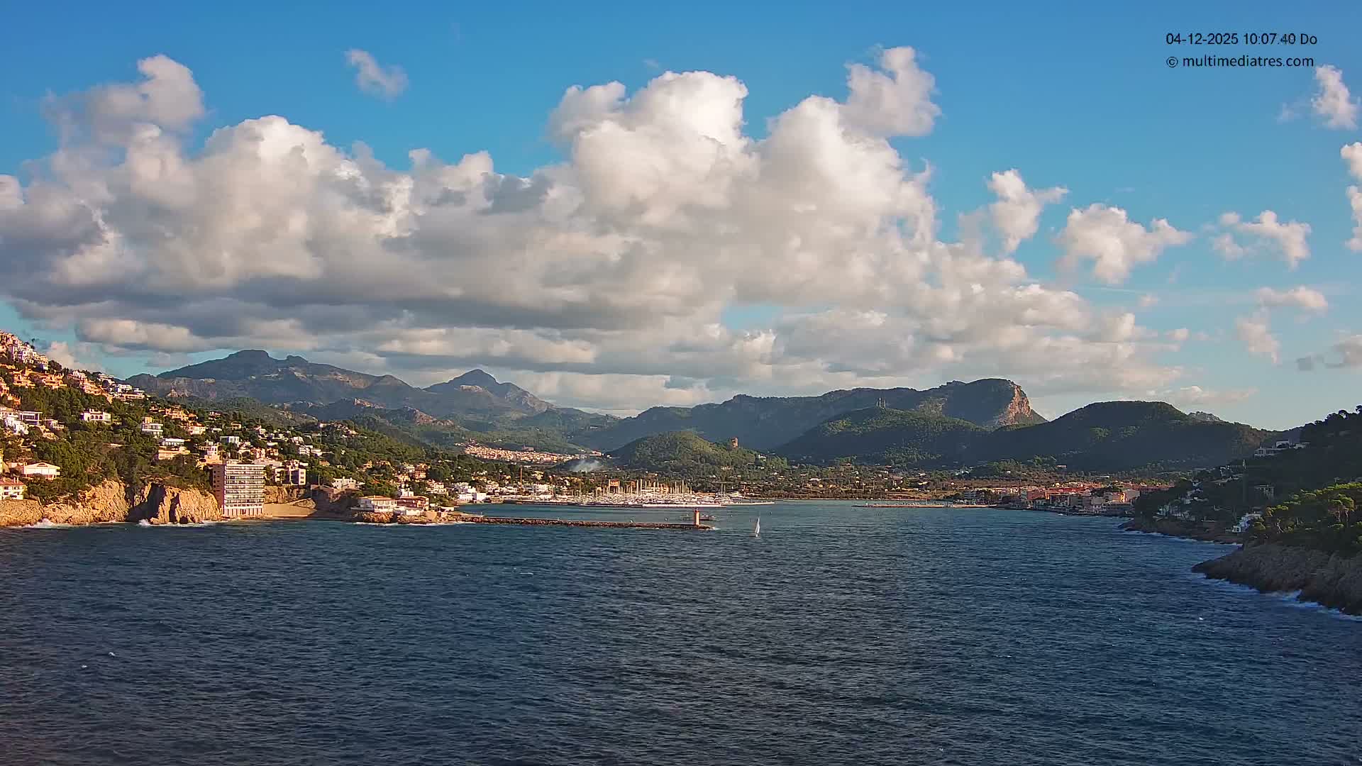 A picturesque coastal town with buildings sprawling up green hillsides lines a deep blue bay featuring a pier with a lighthouse and a marina full of boats, all under a bright blue sky dotted with scattered white clouds.