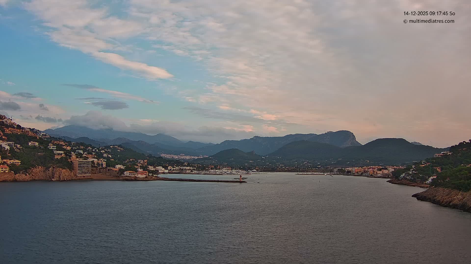 A picturesque coastal town with buildings sprawling up green hillsides lines a deep blue bay featuring a pier with a lighthouse and a marina full of boats, all under a bright blue sky dotted with scattered white clouds.