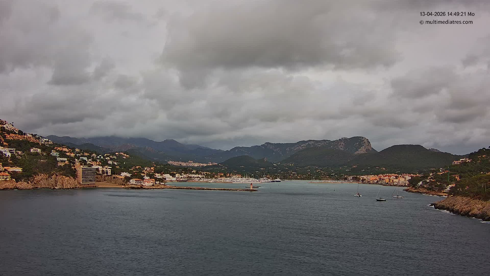A picturesque coastal town with buildings sprawling up green hillsides lines a deep blue bay featuring a pier with a lighthouse and a marina full of boats, all under a bright blue sky dotted with scattered white clouds.