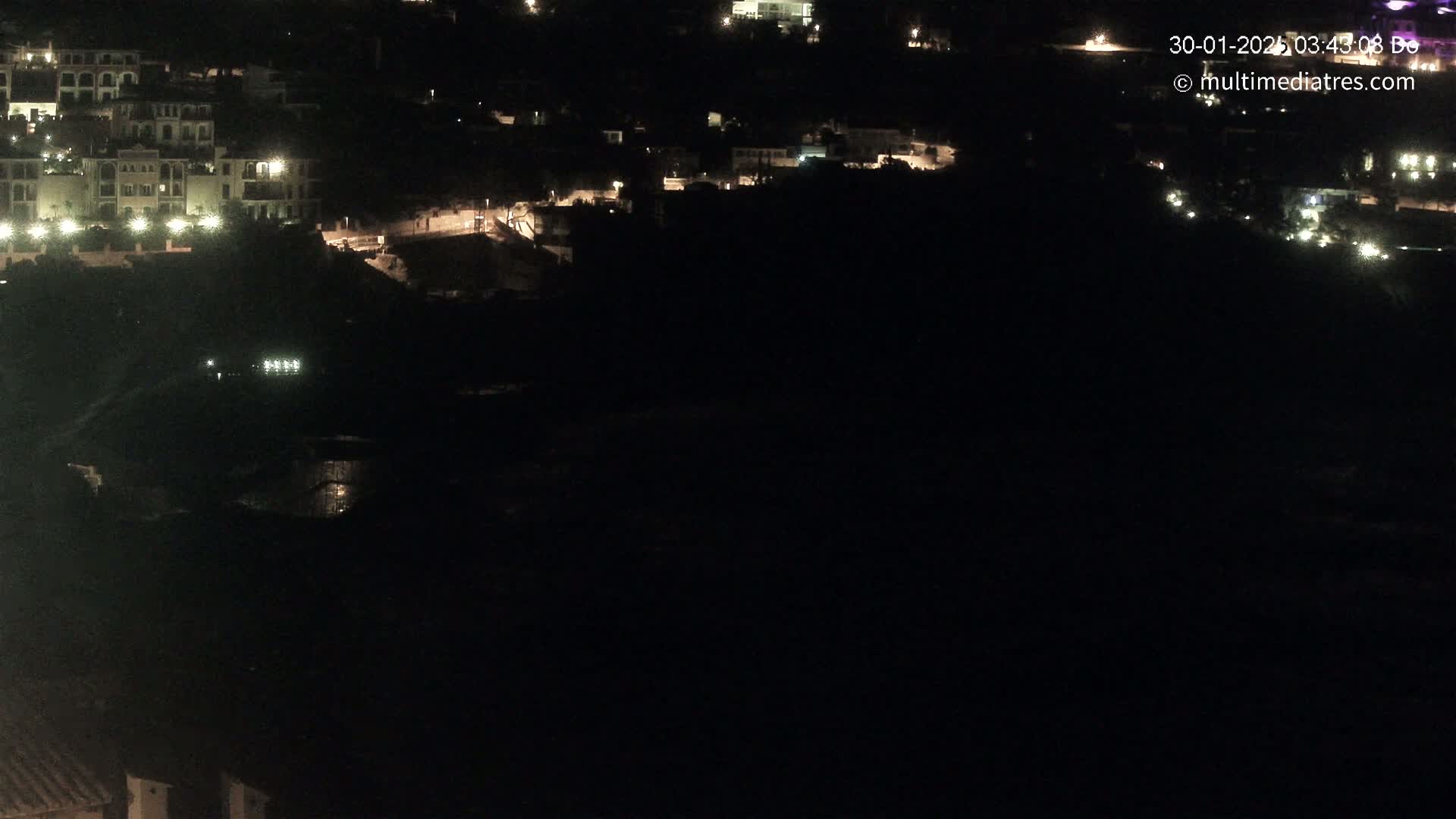 A nighttime aerial view shows a hillside town with sparsely lit buildings and a dark, seemingly calm body of water below.