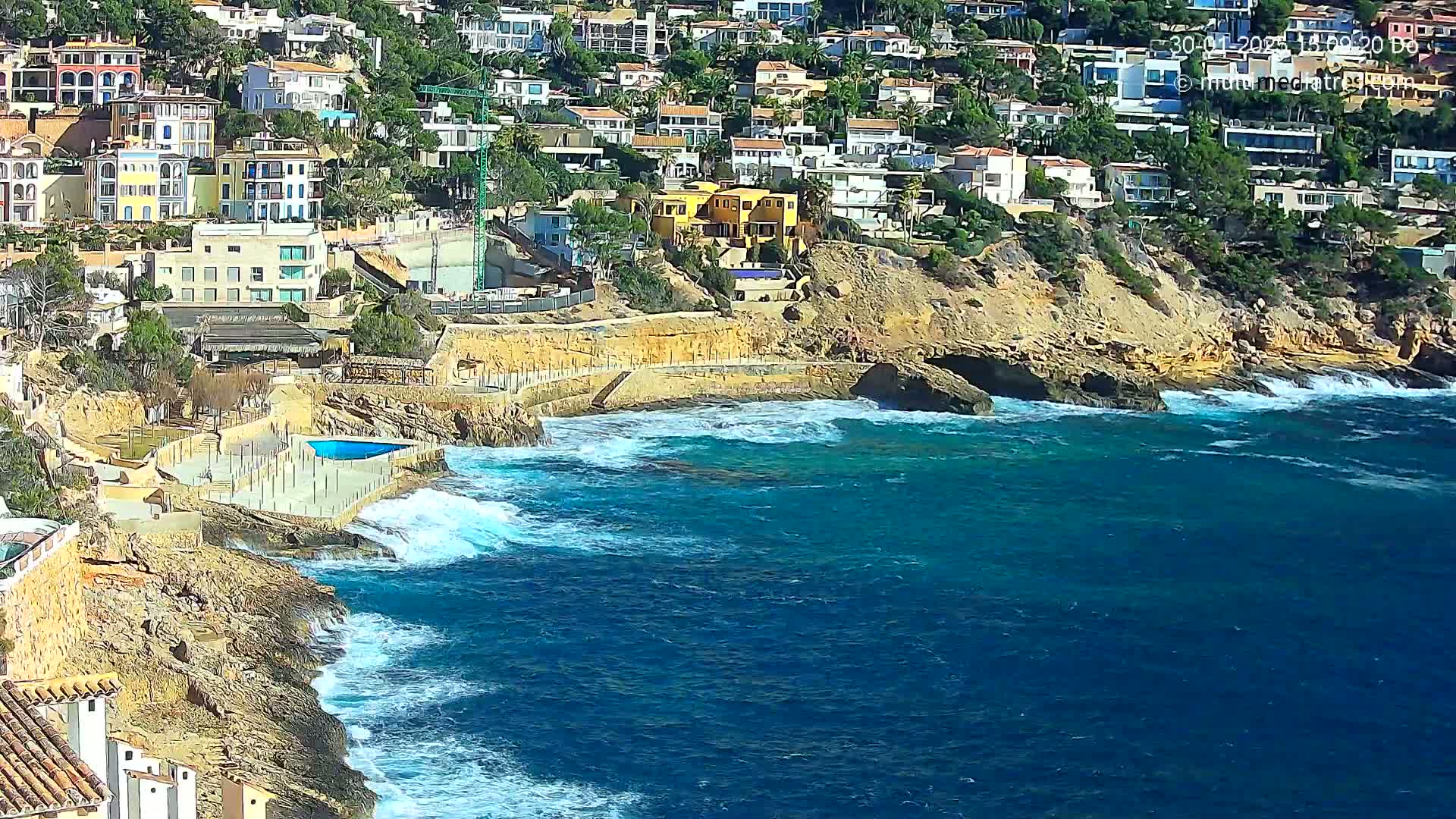 A rocky coastline with a cluster of buildings overlooking a blue sea with moderate waves.