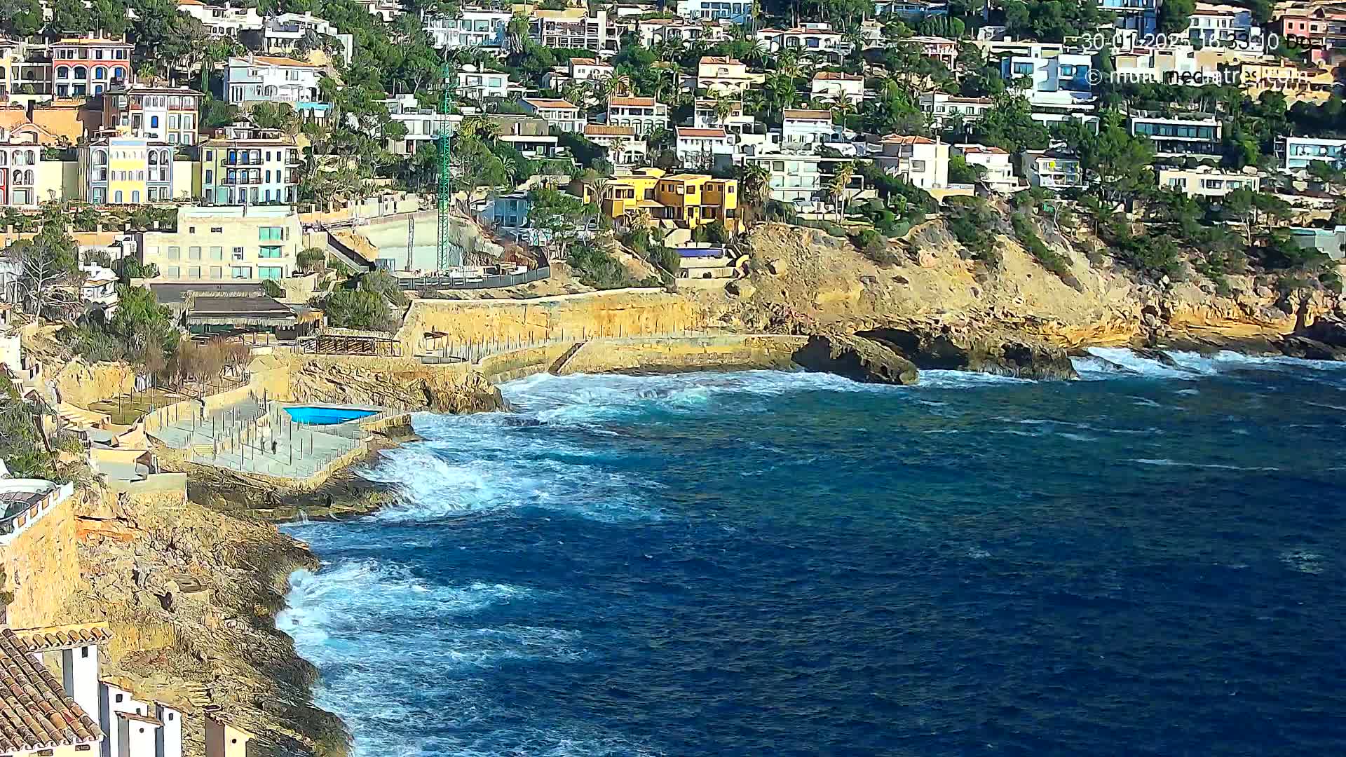 A rocky coastline with a cluster of houses perched on a hillside overlooks a blue sea with moderate waves.