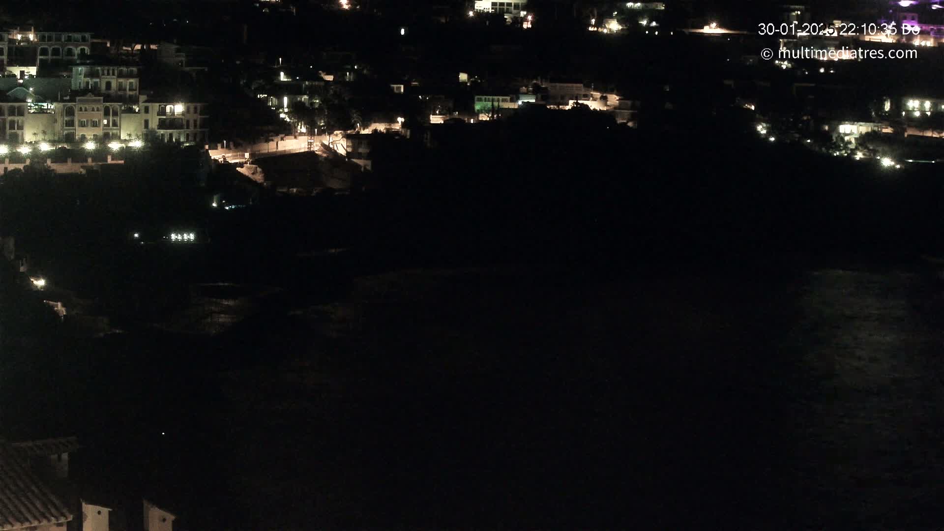 A nighttime, high-angle view shows a dark body of water below a hillside of dimly lit buildings.