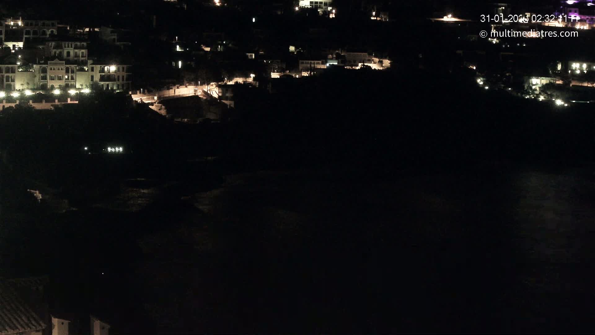 A nighttime, aerial view shows a dark body of water below a hillside of dimly lit buildings.