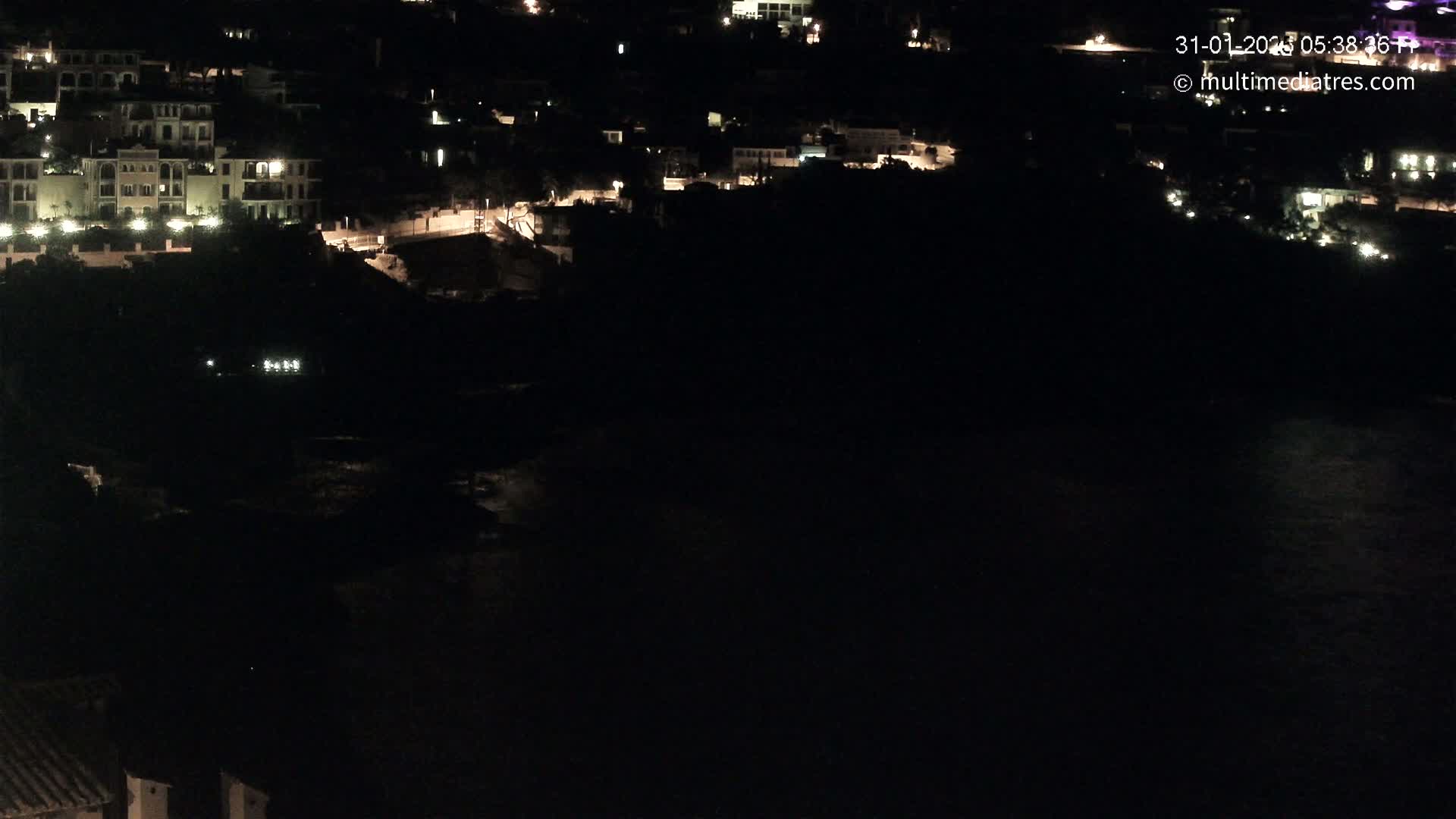 A nighttime, high-angle view shows a coastal town with sparsely lit buildings and a dark body of water below.