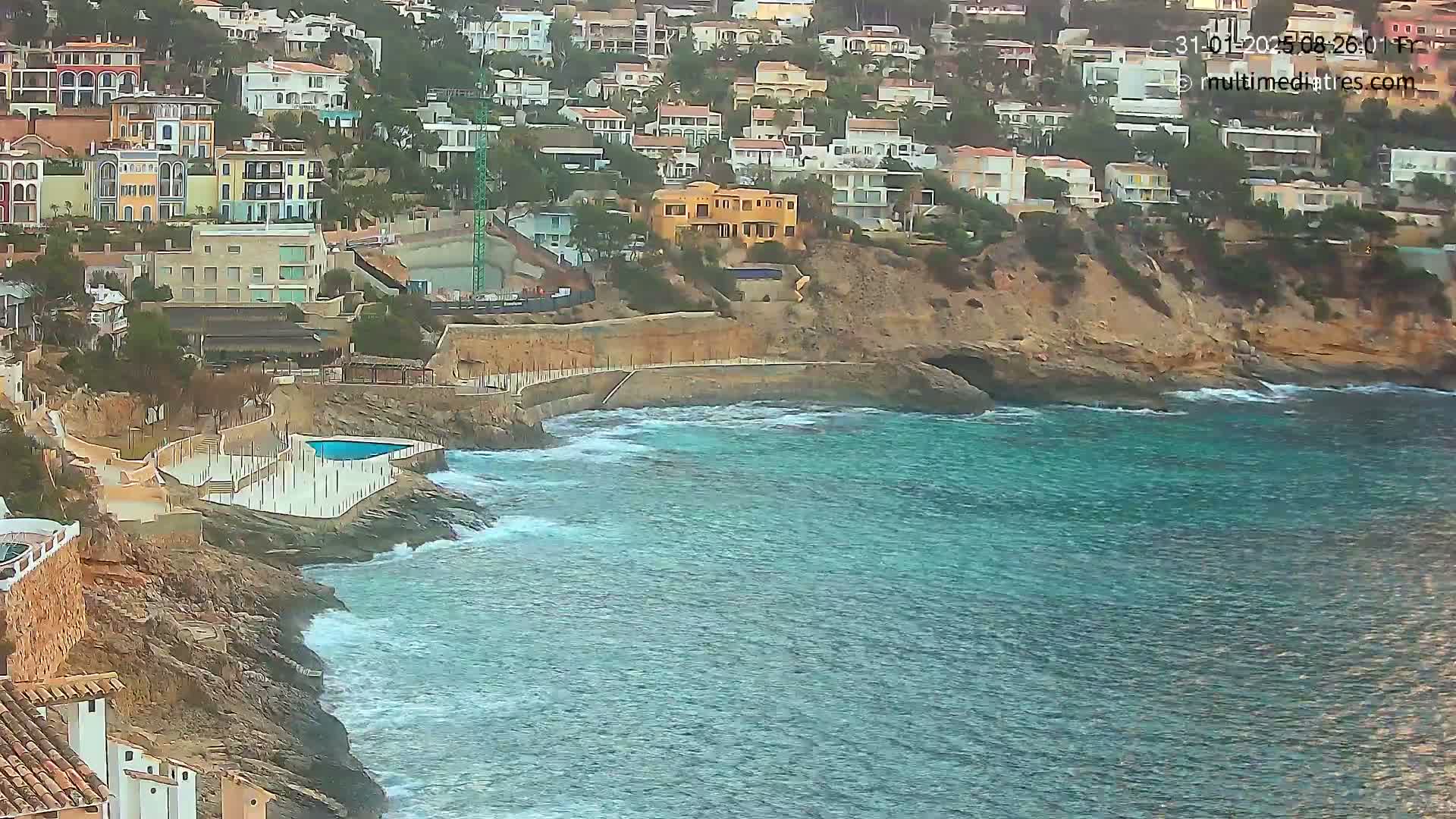 A rocky coastline with colorful houses perched on a hillside overlooks a teal ocean with small waves crashing against the shore.