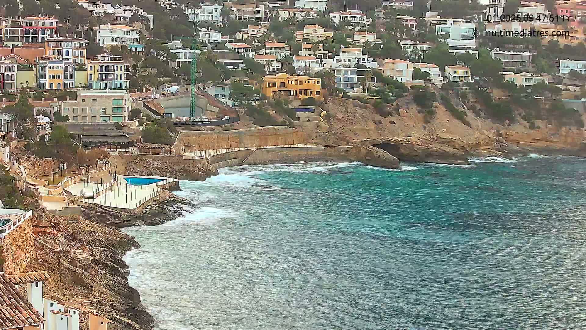 A coastal scene shows a rocky shoreline with buildings perched on a hillside above a small cove with turquoise water and moderate waves.