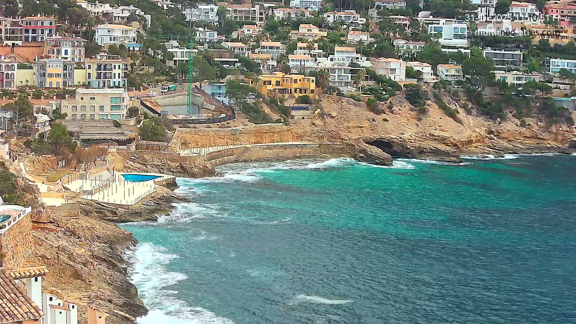 A rocky coastline with teal water and waves laps against the shore near a collection of multi-colored houses built into a hillside.